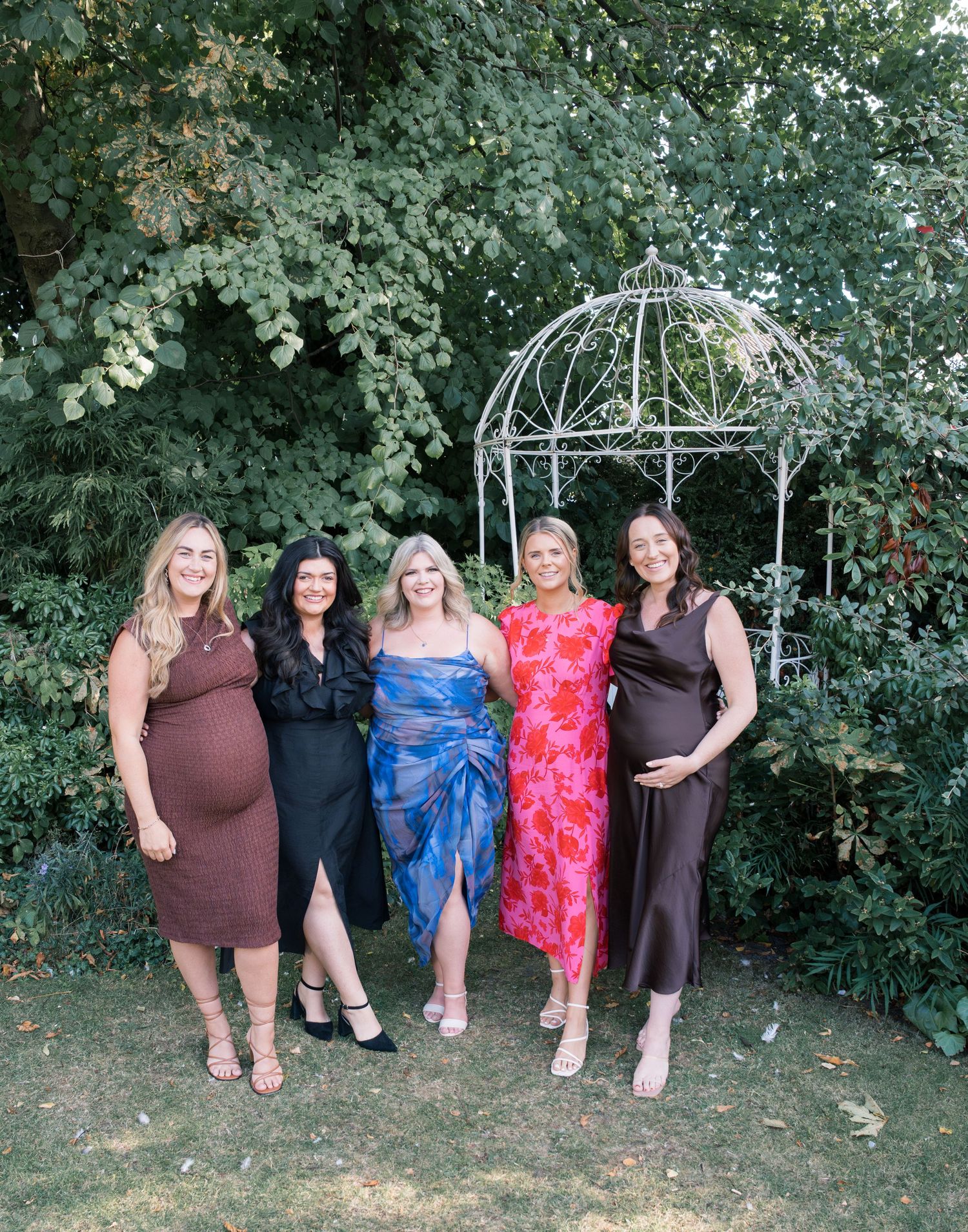 Five women in formal dresses pose in front of white garden gazebo during summer event.