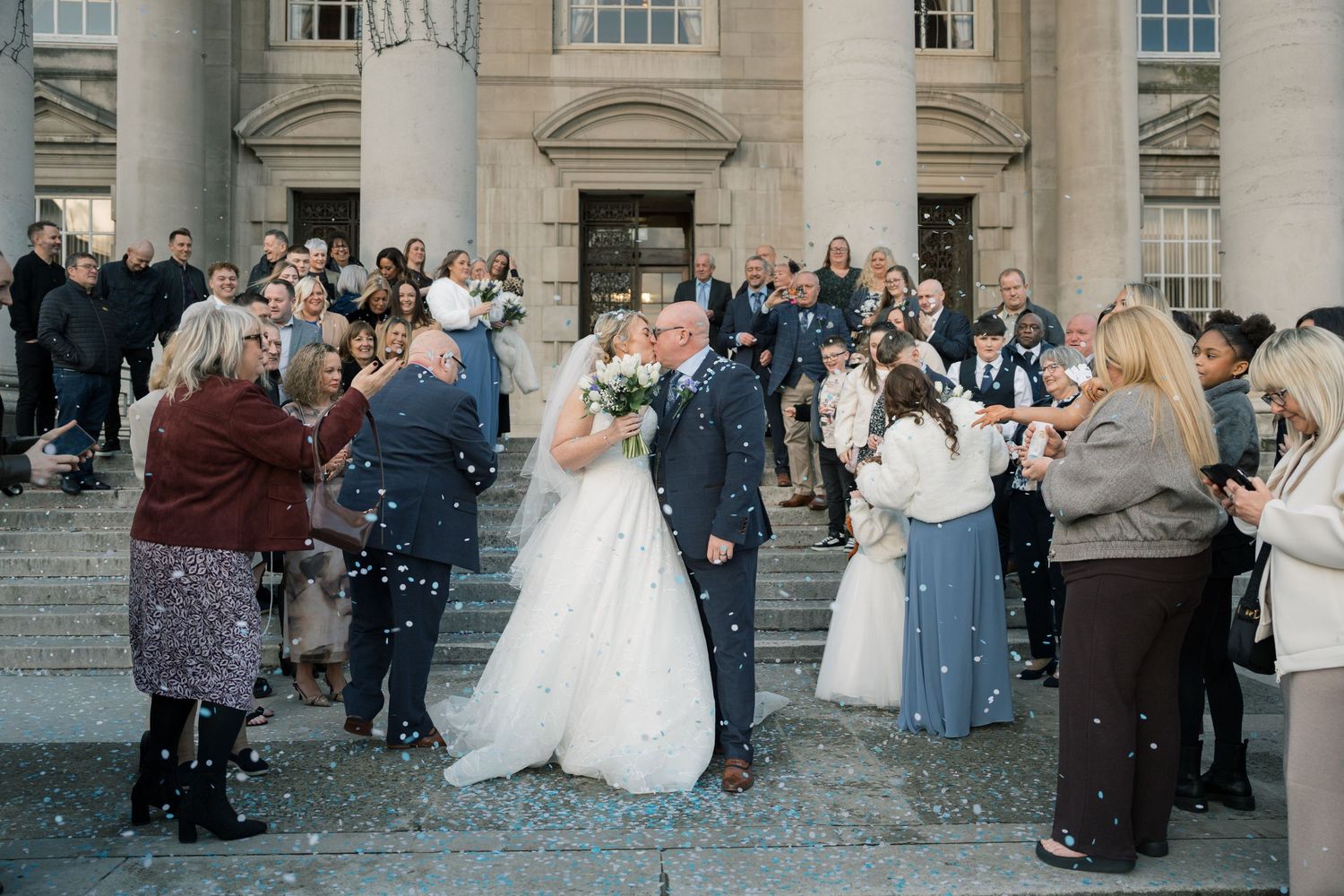 Wedding guests toss flower petals at a couple during their grand exit celebration outside Leeds Civic Hall.