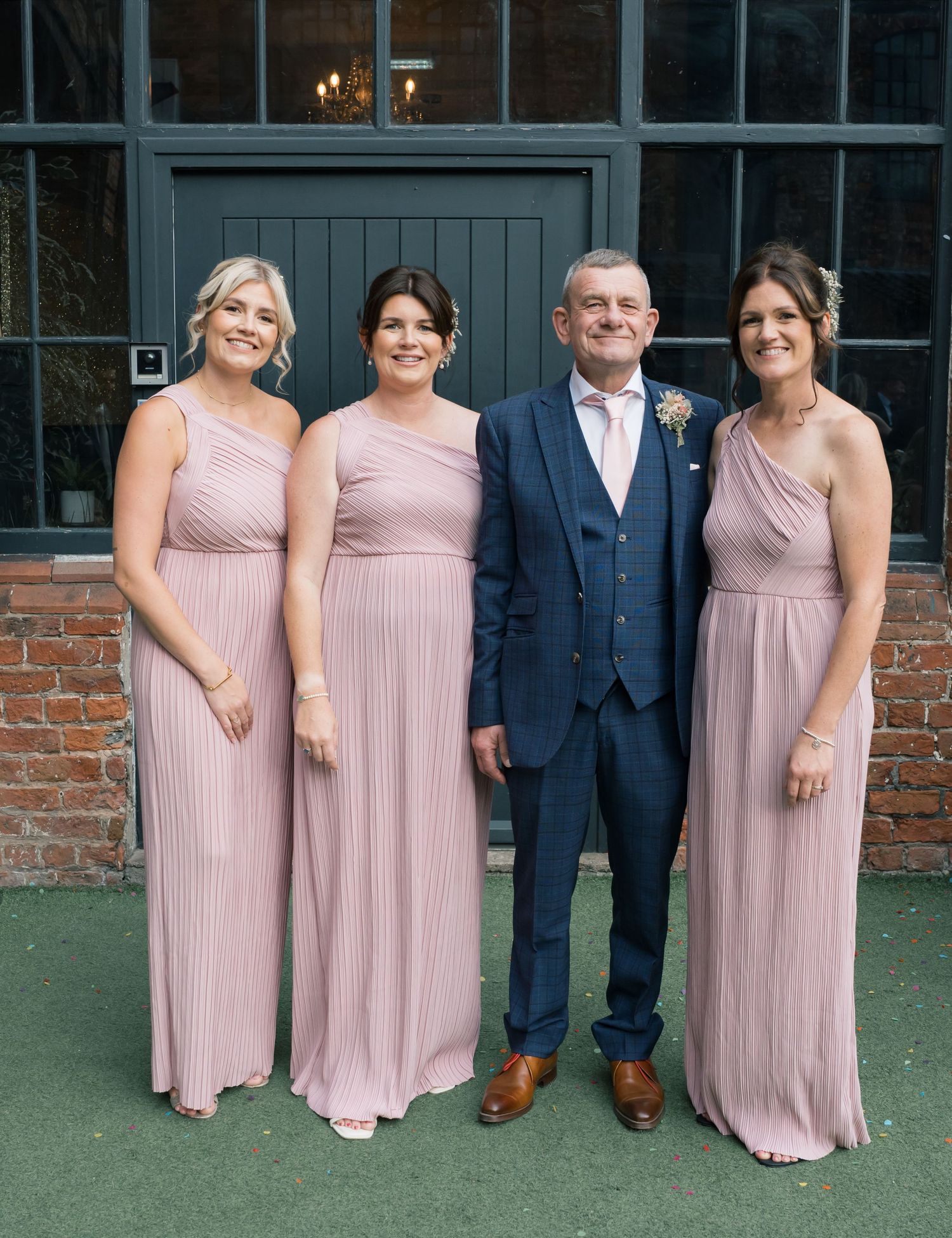 Wedding party members in pink dresses and navy suit pose against brick building.