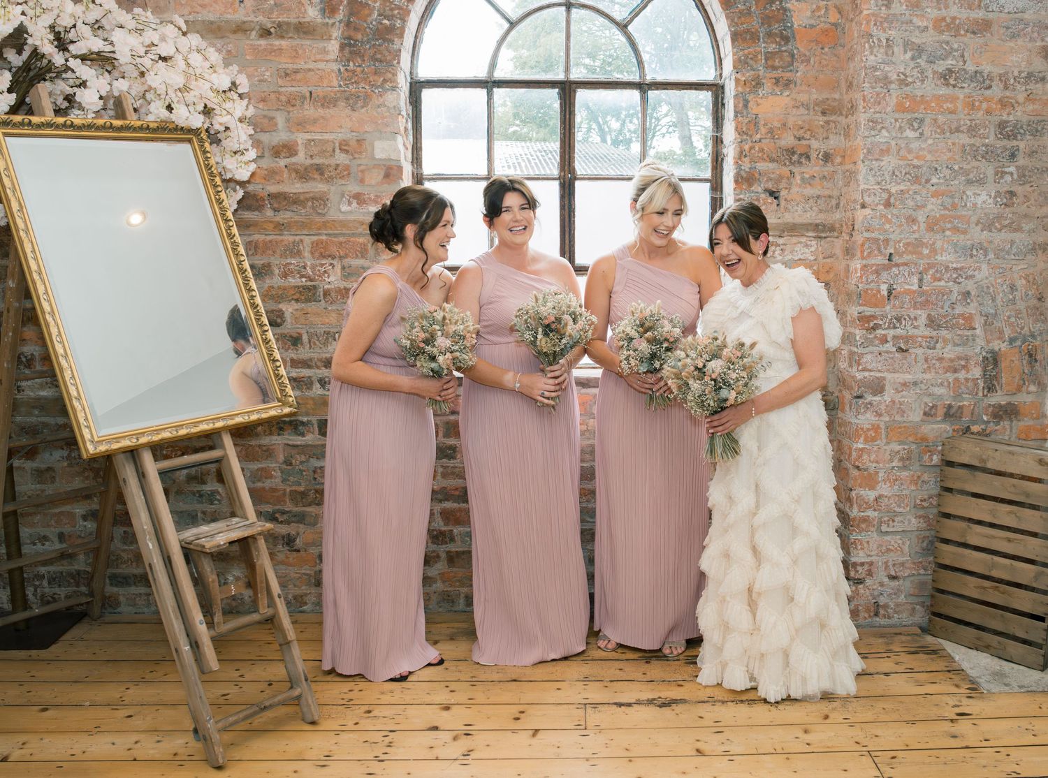 Bridesmaids in dusty pink dresses pose with bride in white gown against rustic brick wall.