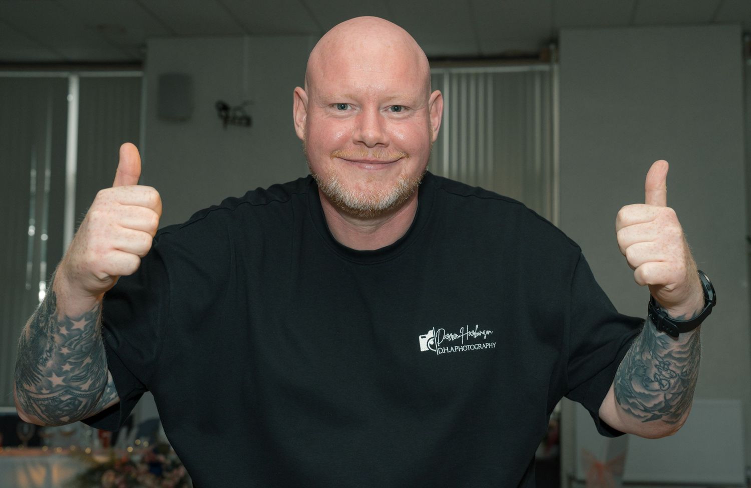 Bald tattooed man in black shirt giving two thumbs up and smiling indoors.
