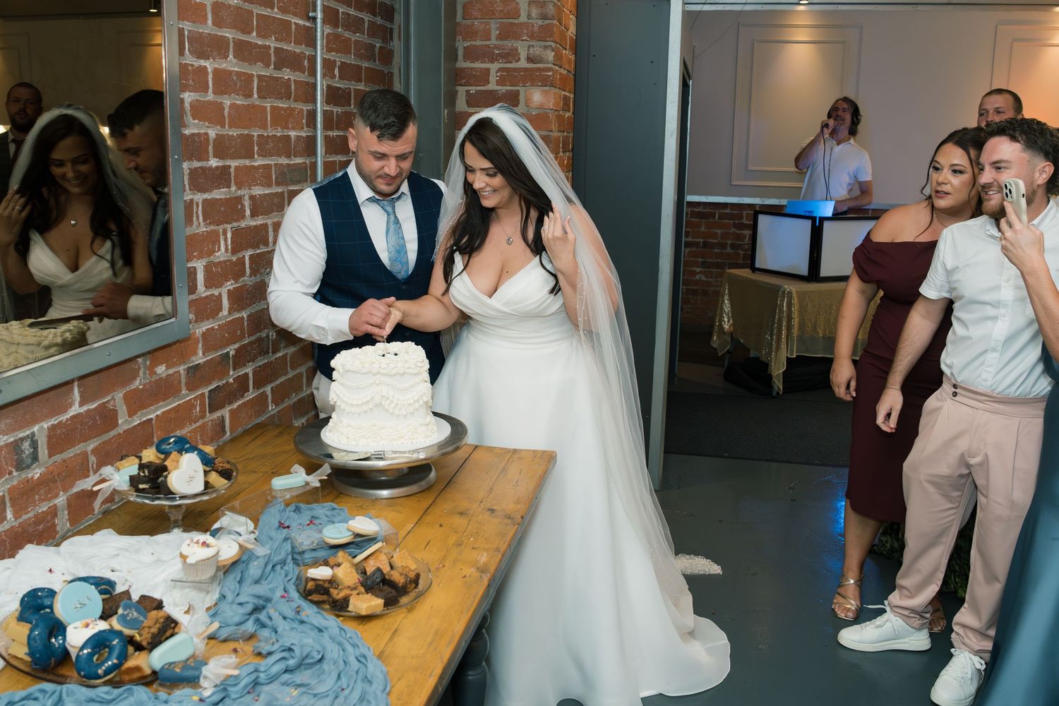 A couple in wedding attire cuts a white and blue cake together at their reception venue with brick walls.