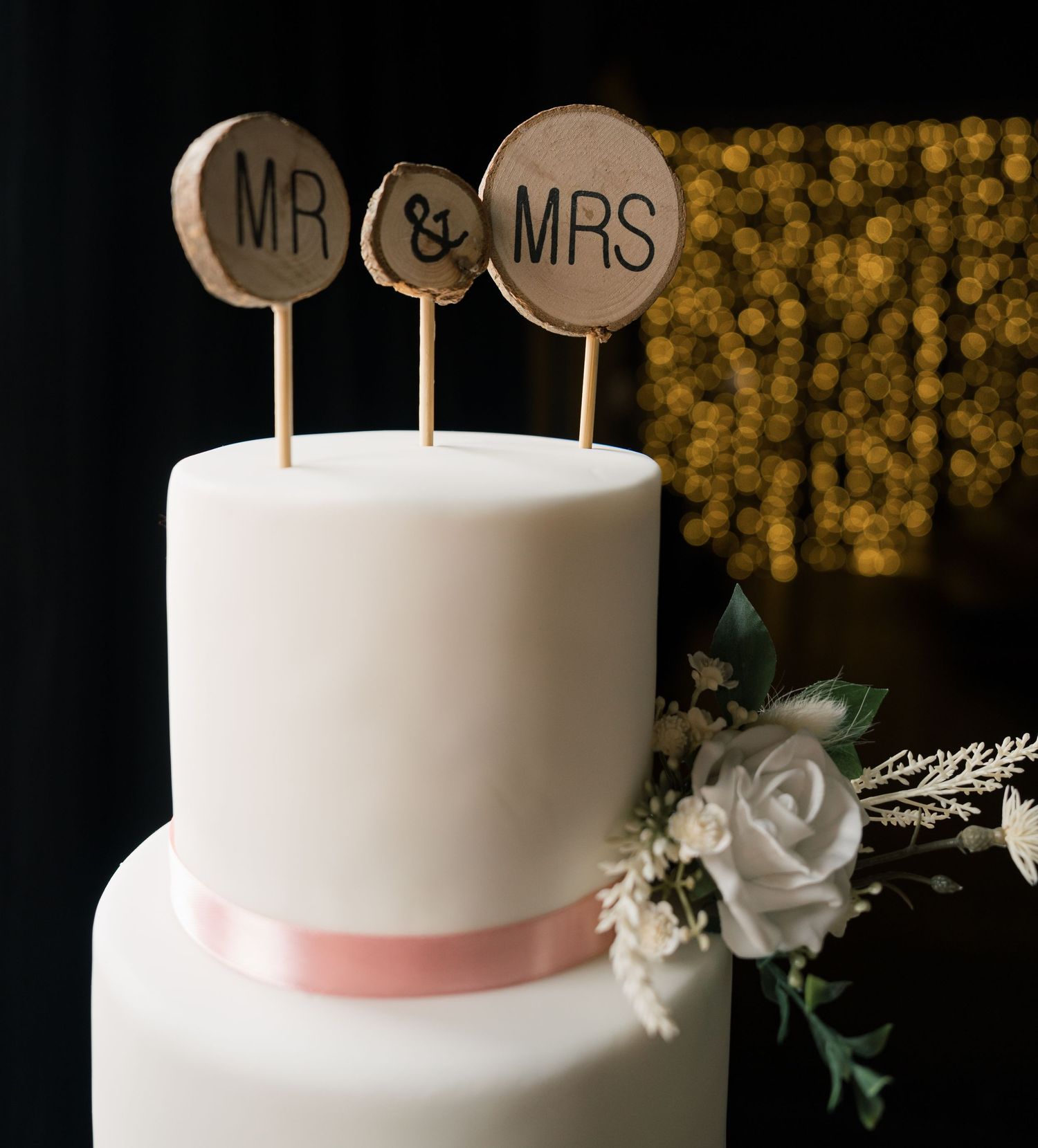 White wedding cake with Mr & Mrs toppers and floral decoration against a bokeh background.