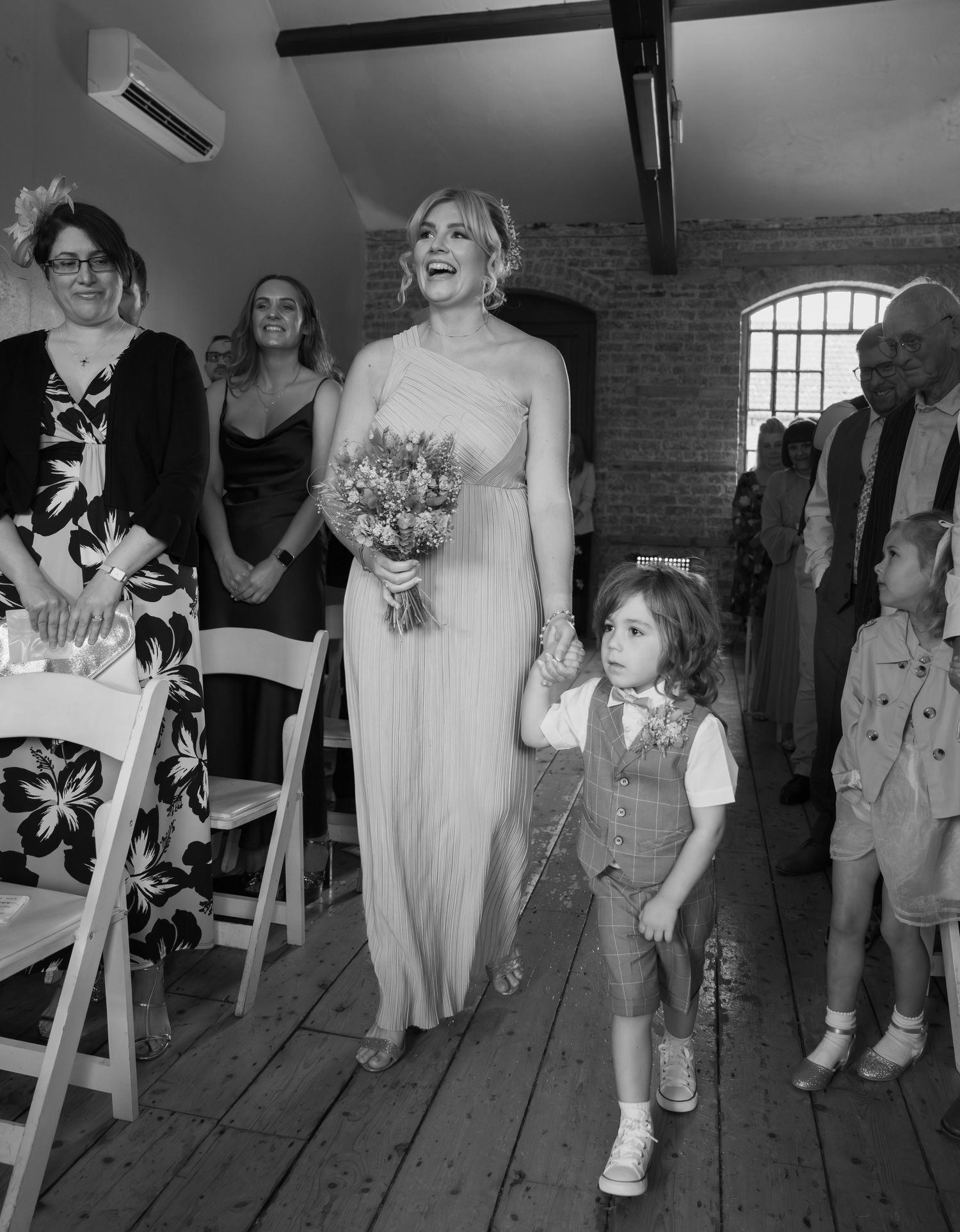 Black and white photo of wedding guests mingling in a rustic brick venue with wooden floors.