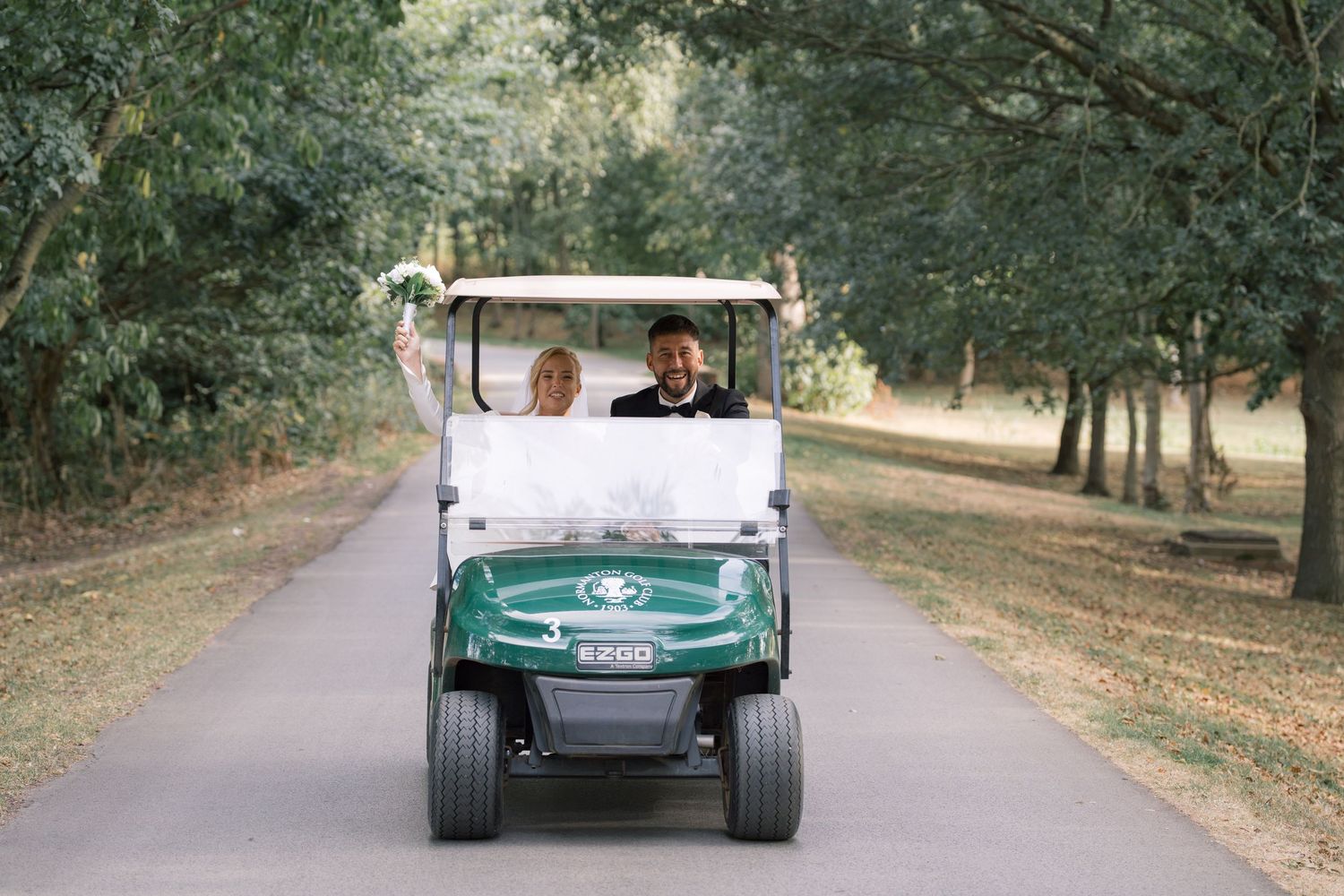Bride and groom riding a green golf cart down a tree-lined path, celebrating their wedding day outdoors.