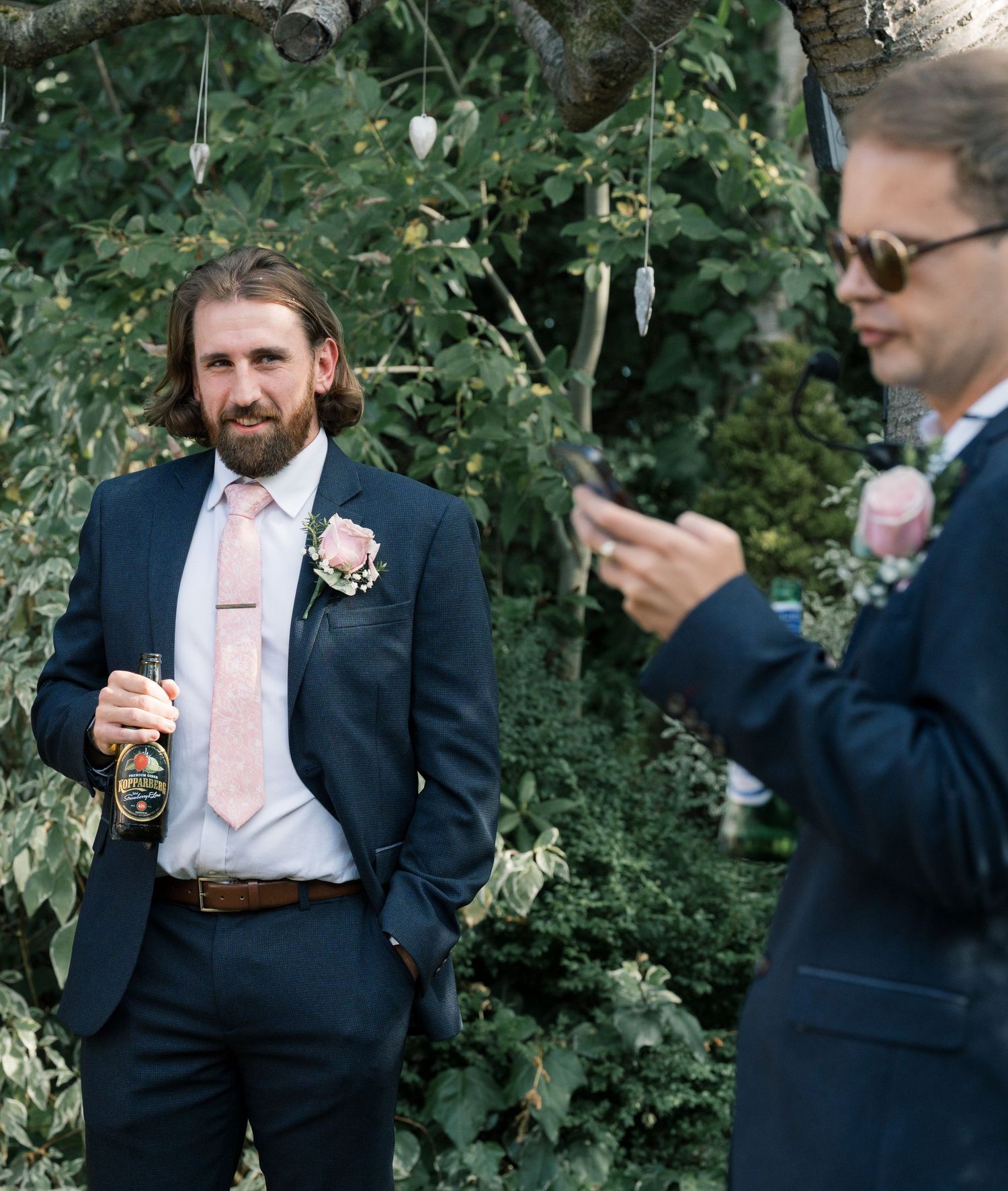 Groomsmen in navy suits with boutonnieres enjoying drinks during an outdoor wedding celebration.