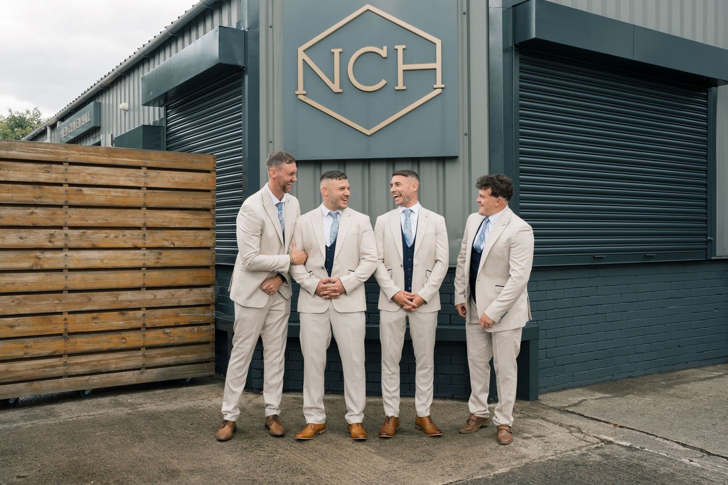 A group in light gray suits sit casually on wooden benches against an industrial metal building exterior.