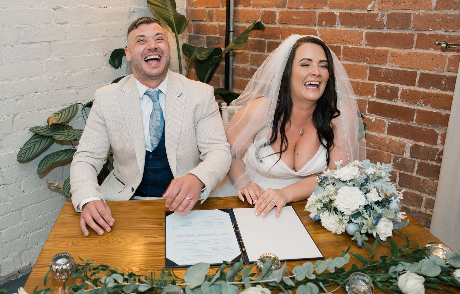 Couple shares joyful laughter while seated at rustic wooden table with exposed brick wall and greenery decorations.