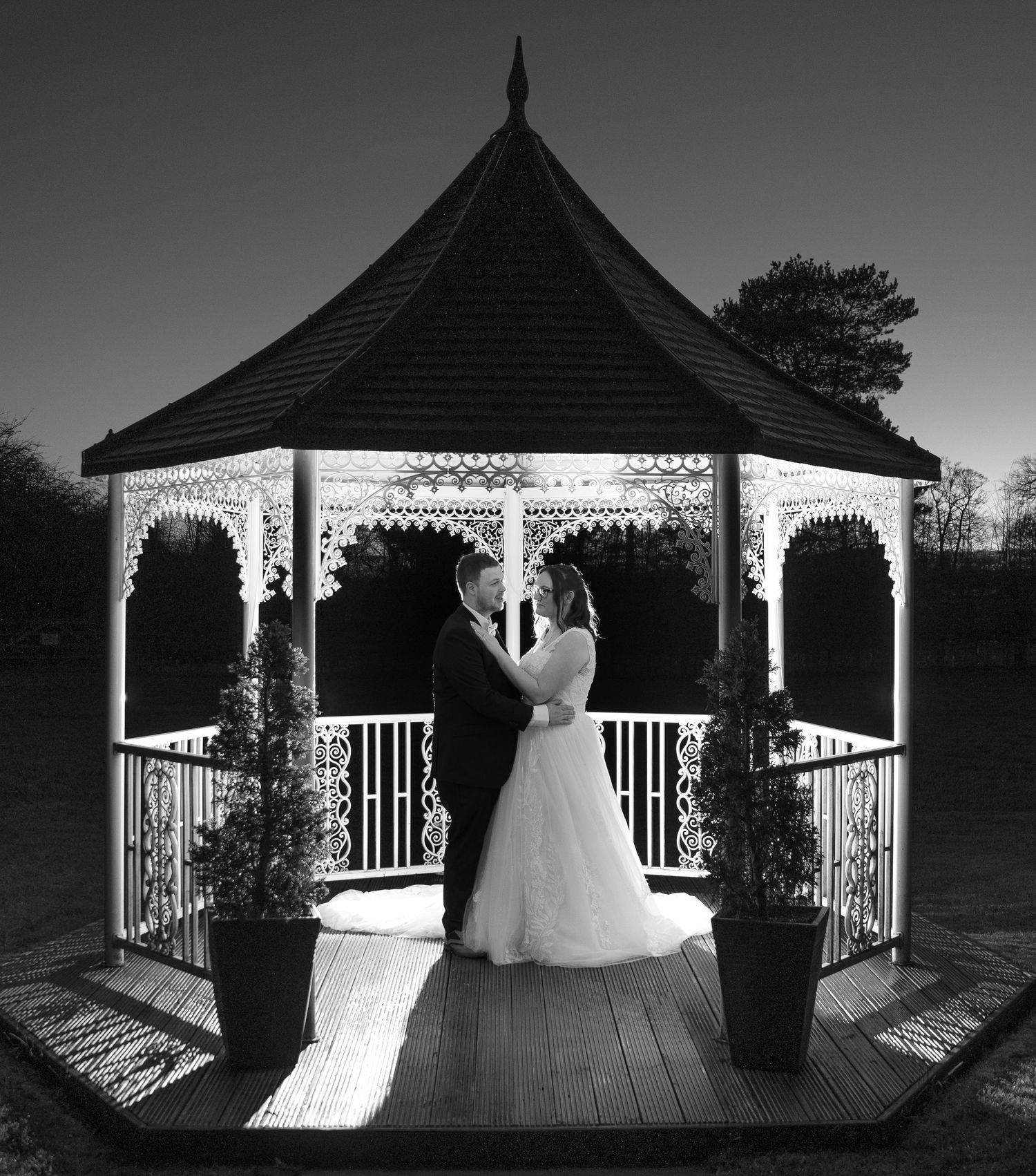 A black and white photograph of a couple in an ornate Victorian-style gazebo at dusk with decorative potted plants.