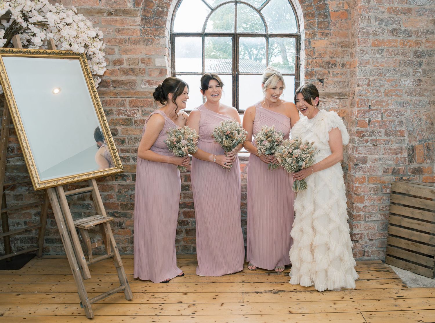 A wedding party in dusty pink bridesmaid dresses poses together in a rustic brick venue with an elegant mirror display.