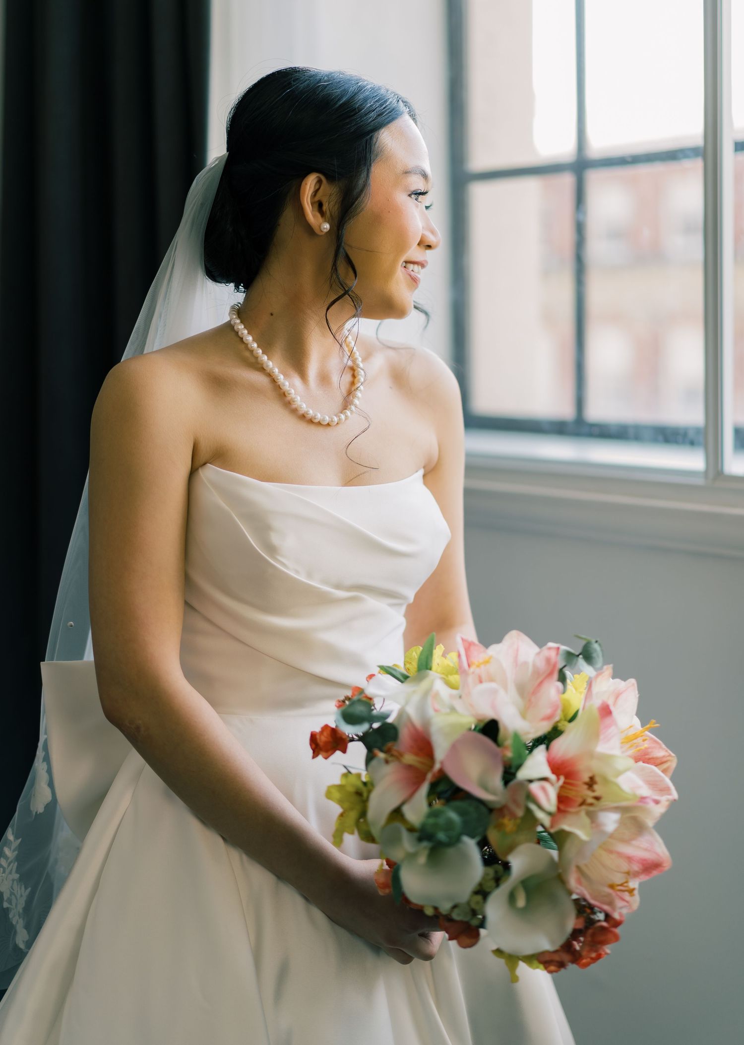 A bride in a white strapless wedding dress holds a pink and white floral bouquet while looking out a sunlit window.