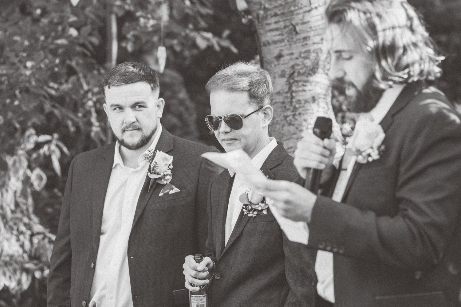 Wedding guests in formal attire gathered outdoors during a ceremony with one speaking into a microphone.