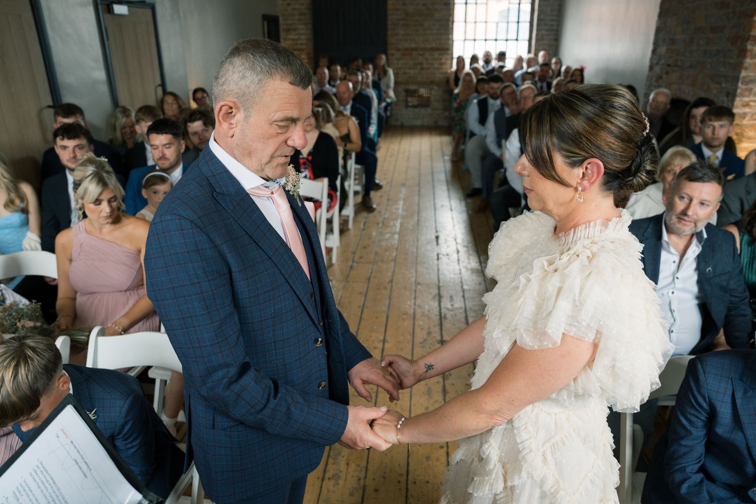 Newlyweds share their first dance surrounded by guests in a rustic brick-walled venue with natural lighting.