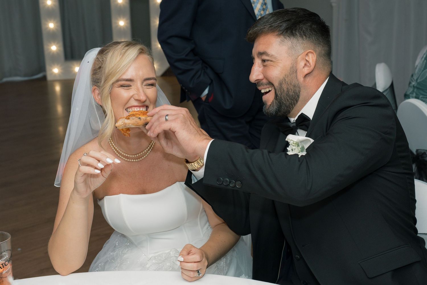 Bride and groom laughing while sharing food at wedding reception, bride in white dress, groom in dark suit with boutonniere.