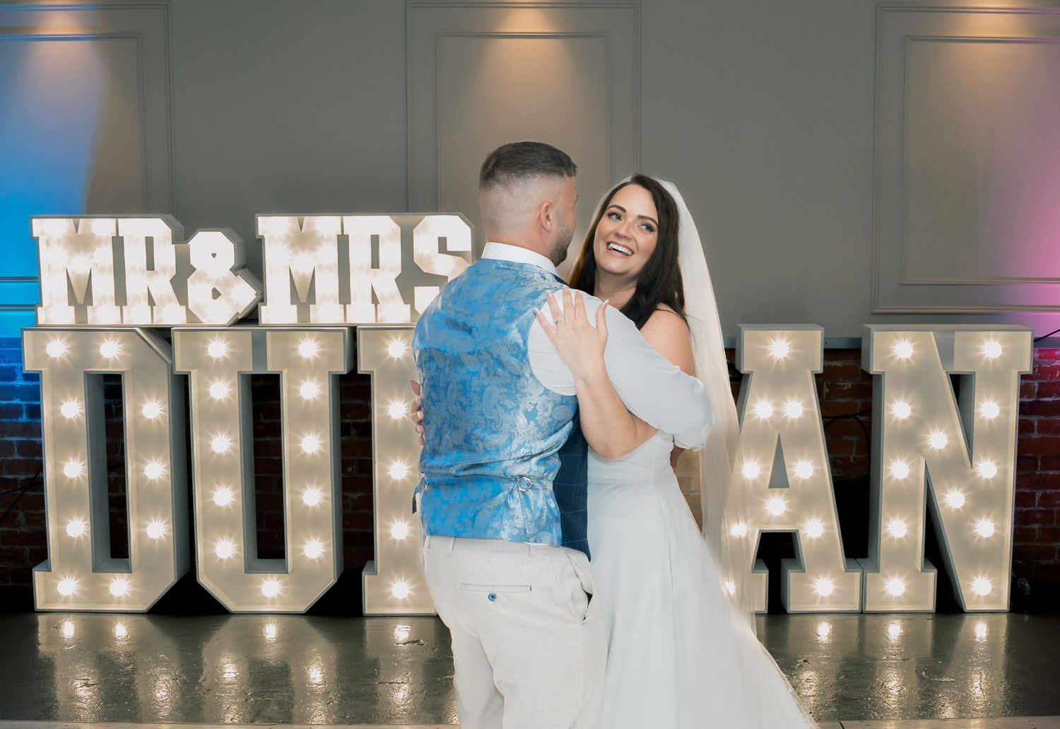 A bride and groom dance in front of illuminated 'MR & MRS' marquee letters at their wedding reception.