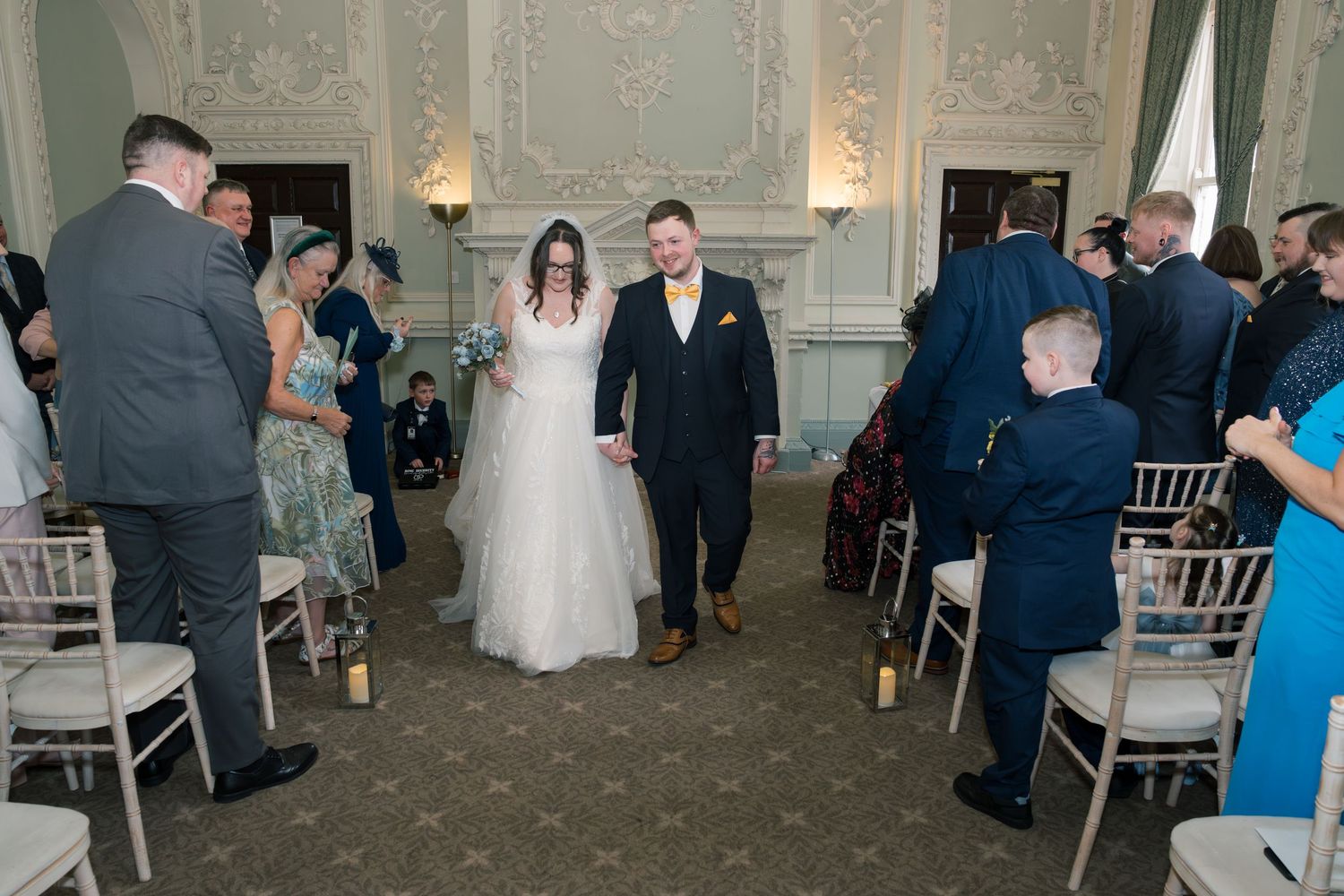 Wedding reception guests mingle and dance in an elegant ballroom with ornate white walls and crystal chandeliers.