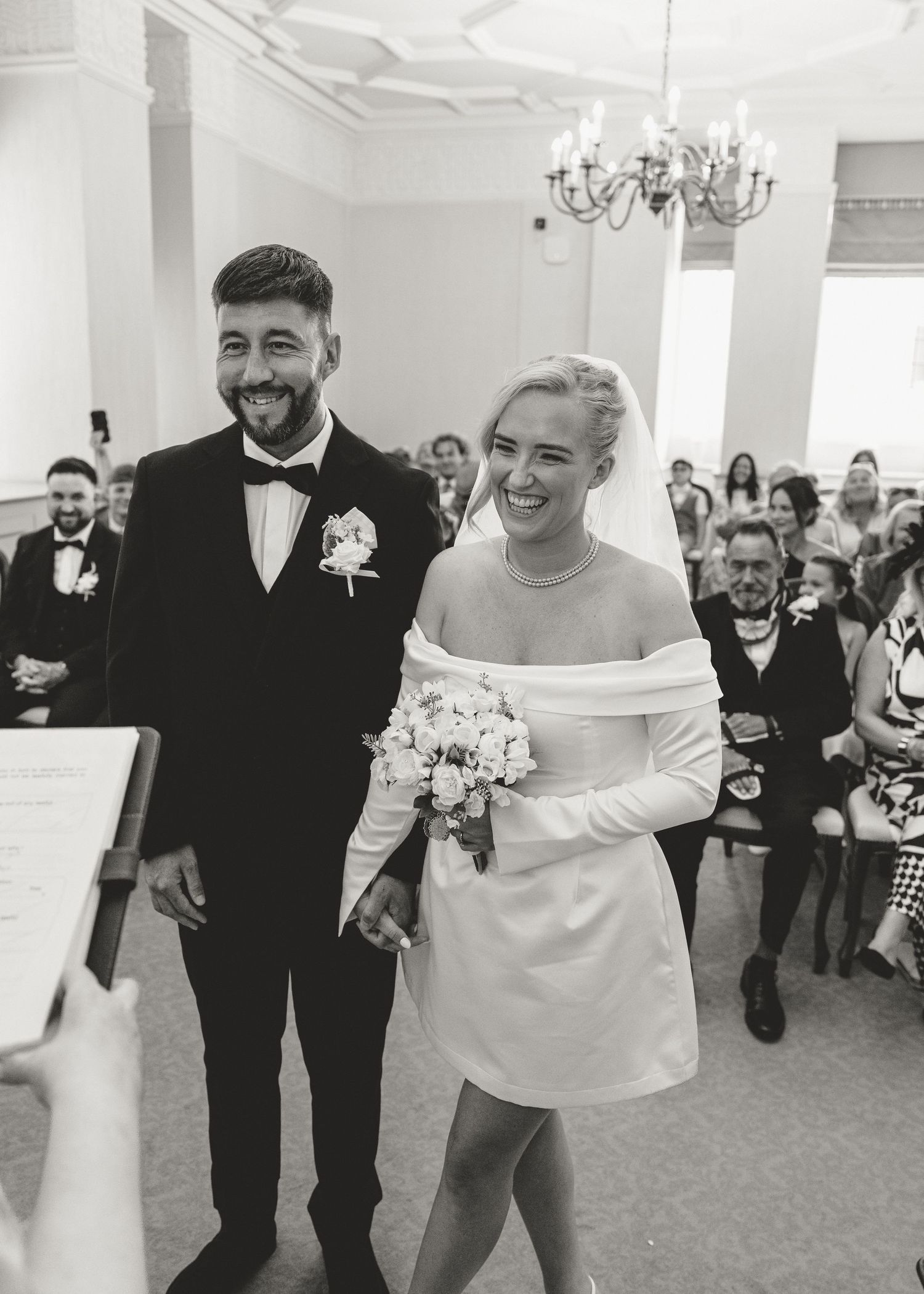 Black and white photo of smiling bride in short white dress holding bouquet and groom in tuxedo at wedding ceremony.