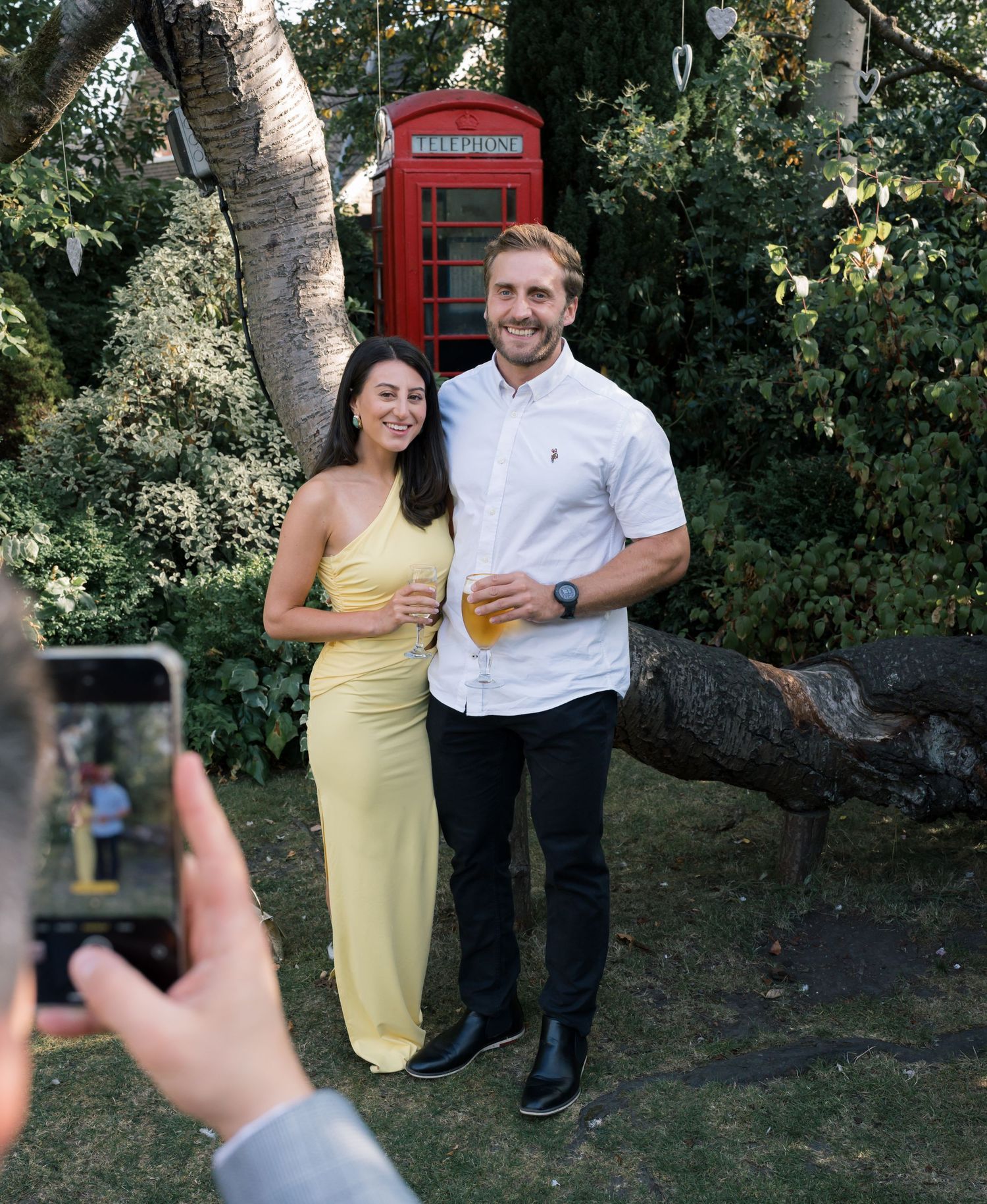 A classic red British phone booth stands among trees in a garden setting.