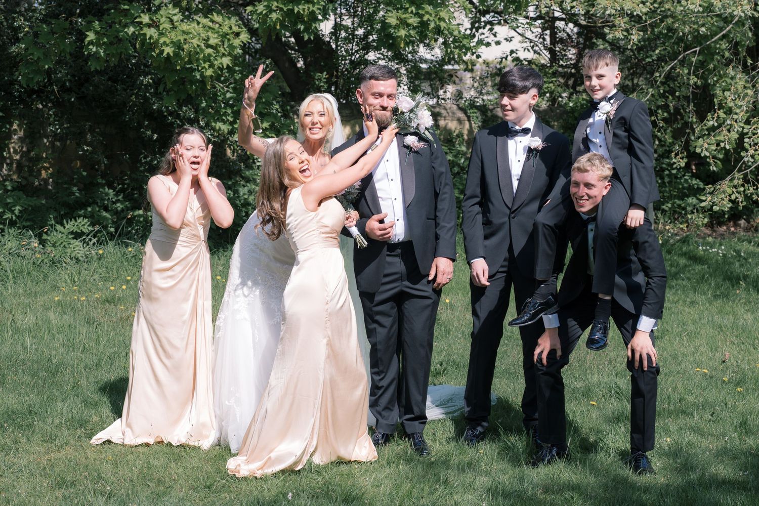 Joyful wedding party celebrating outdoors, bride and groom with bridesmaids and groomsmen laughing on green lawn.