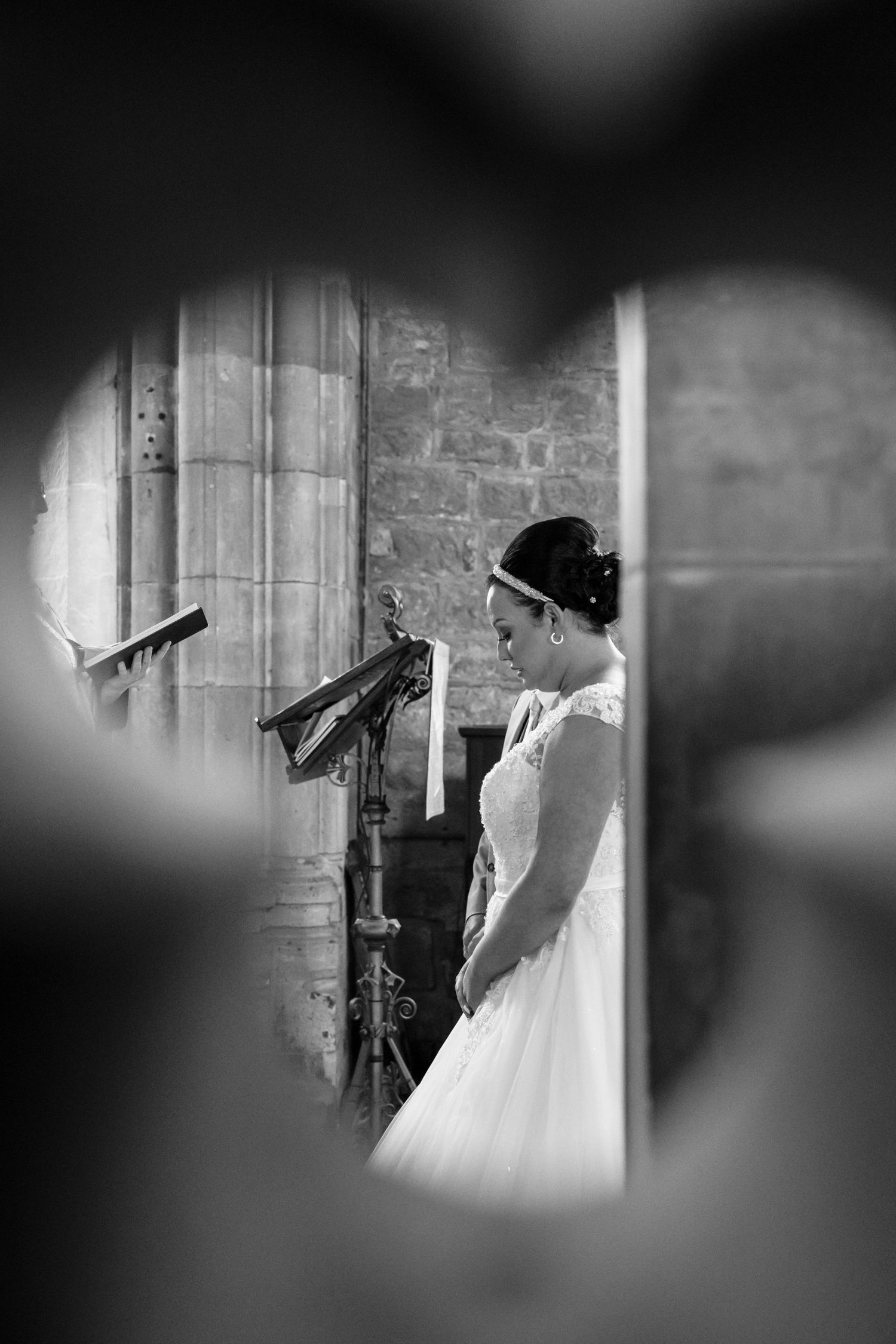 Black and white artistic wedding photograph showing a bride in a white gown standing near a stone church pillar.