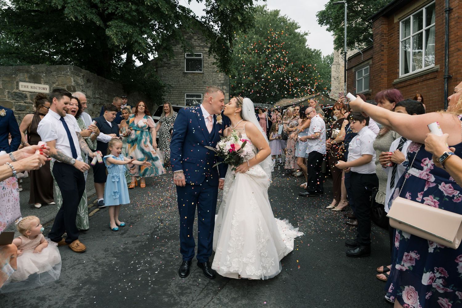 Wedding guests shower the newlyweds with confetti as they share a romantic kiss outside on a summer day.
