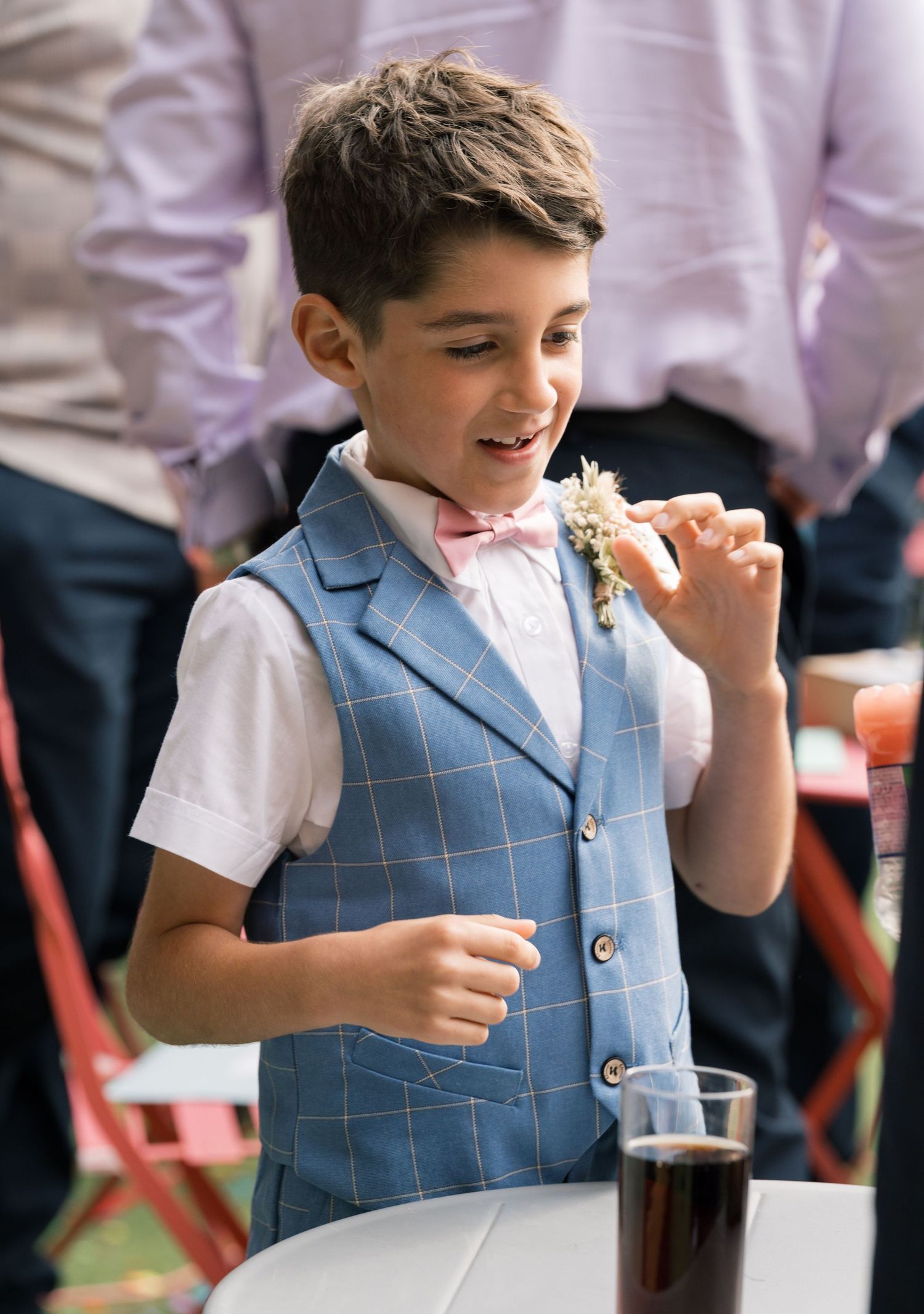 Young boy in light blue checkered vest enjoying a beverage at wedding reception.