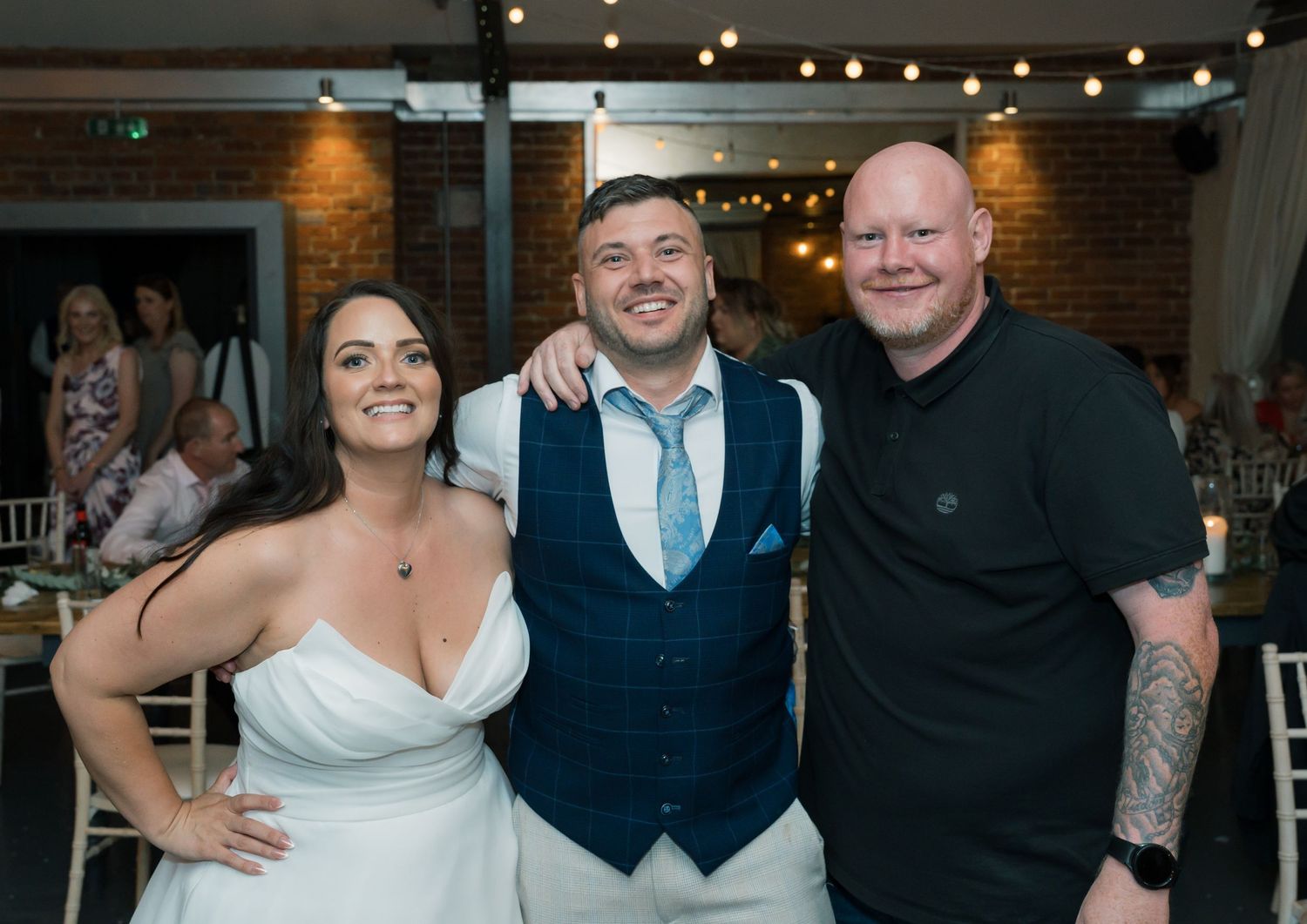 Wedding celebration group poses together inside venue with exposed brick and string lights.