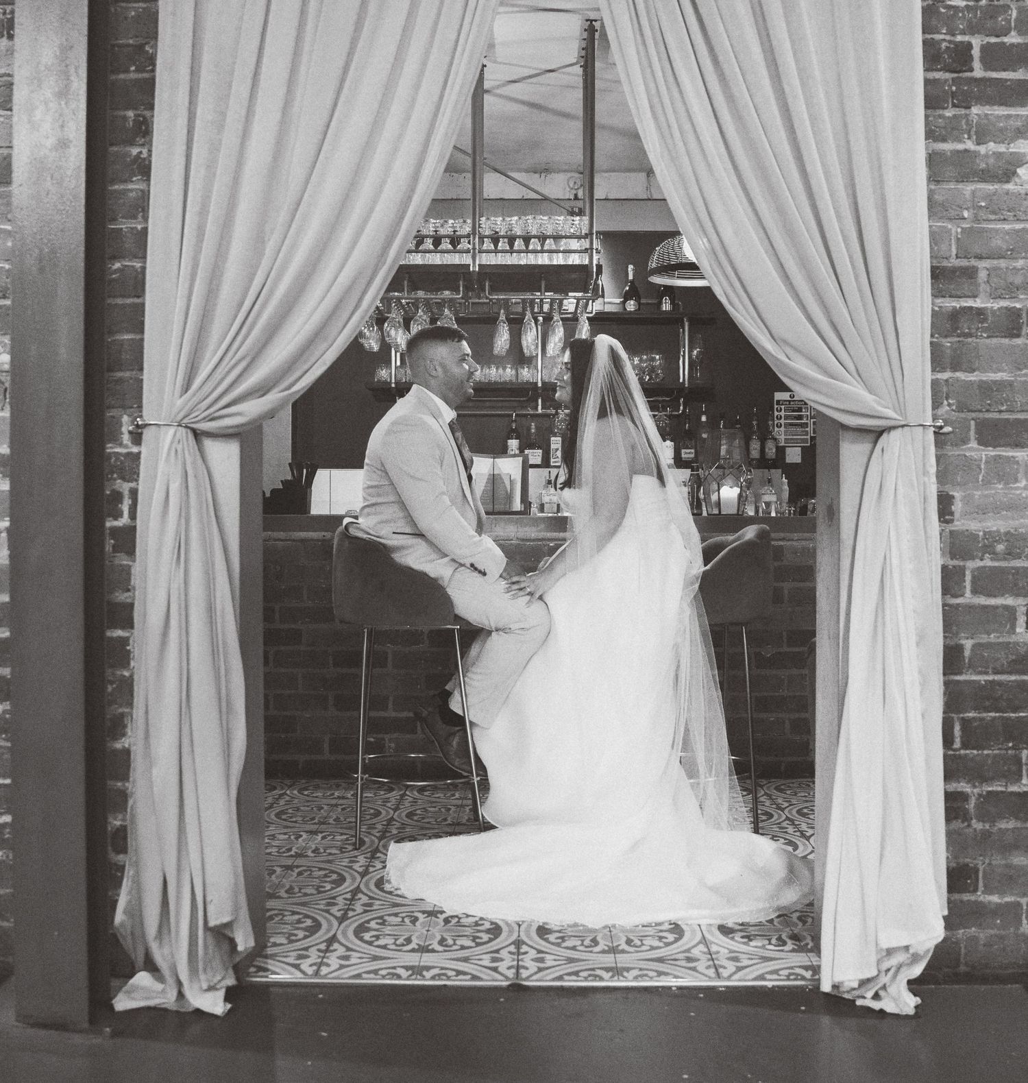 A bride in a white wedding dress poses elegantly against a rustic brick wall with flowing curtains in the background.