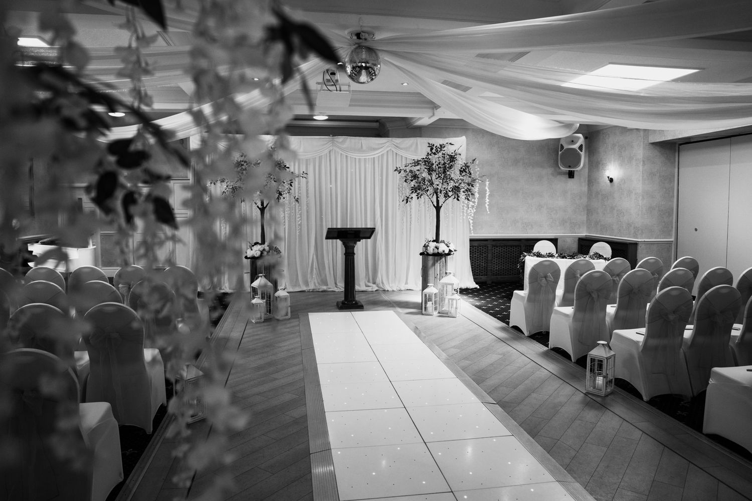 Black and white photo of an indoor wedding ceremony setup with rows of chairs and floral decorations.