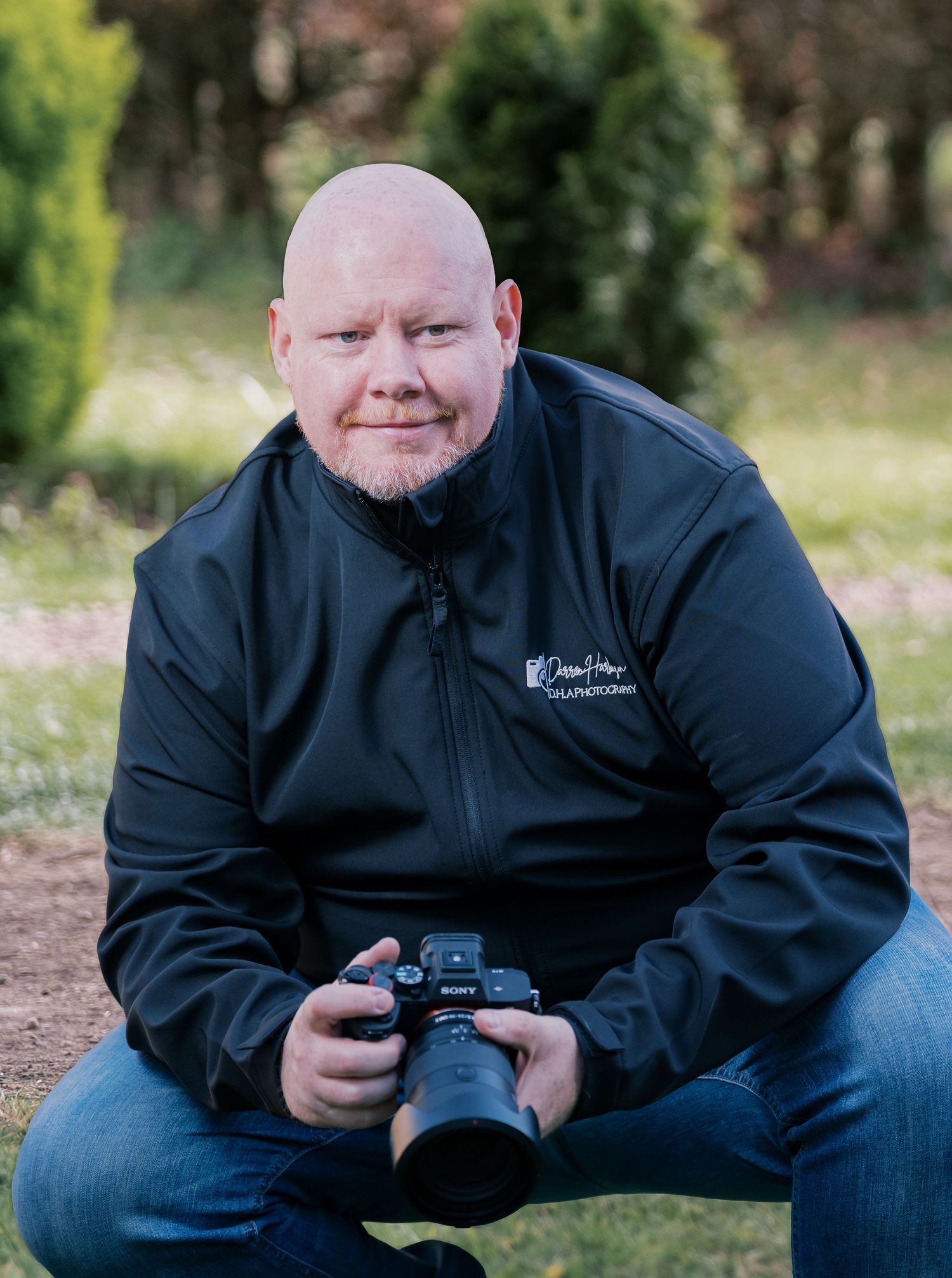A person in a black jacket sits outdoors holding a camera while taking photos in a natural setting.