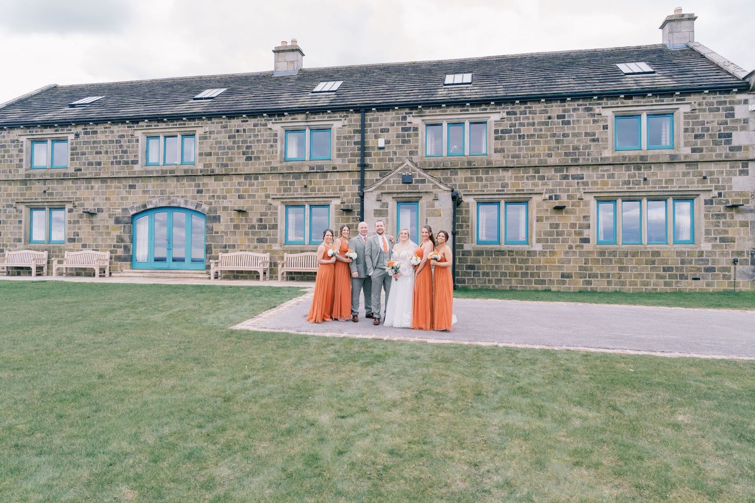 Wedding party in coral dresses poses outside stone building with blue windows.