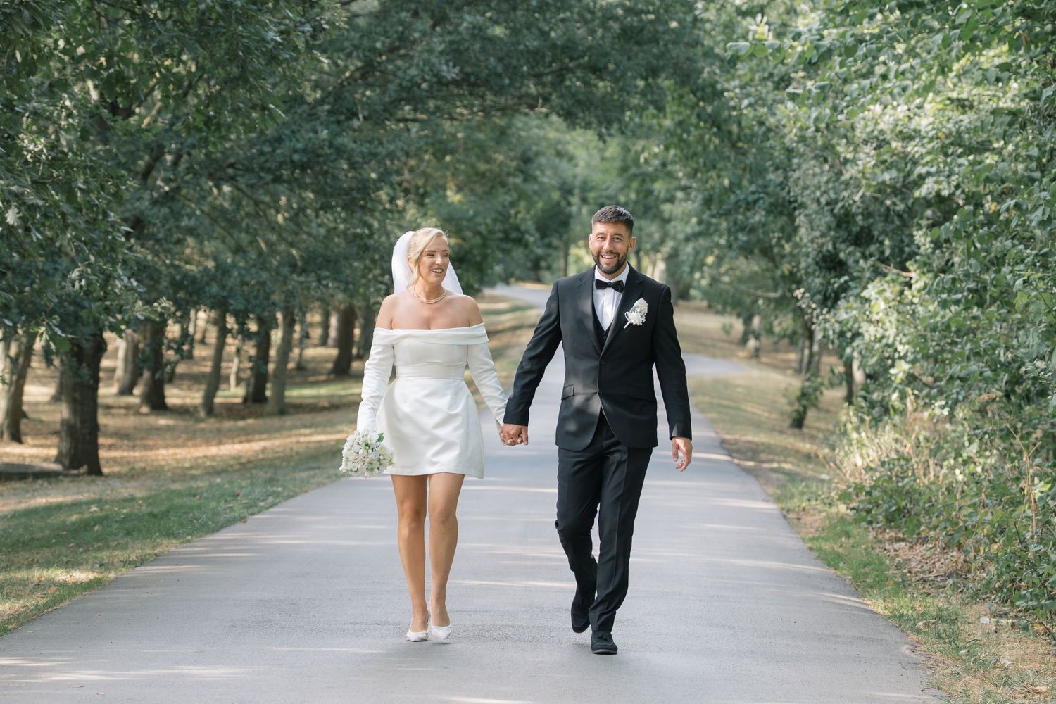 Bride in short white off-shoulder dress and groom in black tuxedo walk hand-in-hand down a tree-lined path.