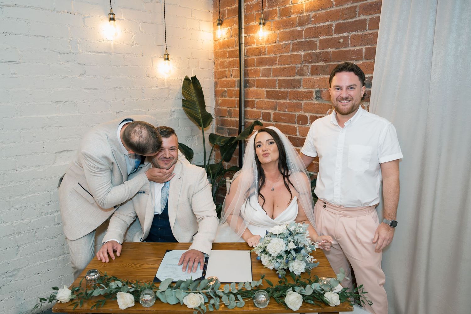 Wedding party gathered in an industrial brick-walled venue with hanging lights and casual atmosphere.