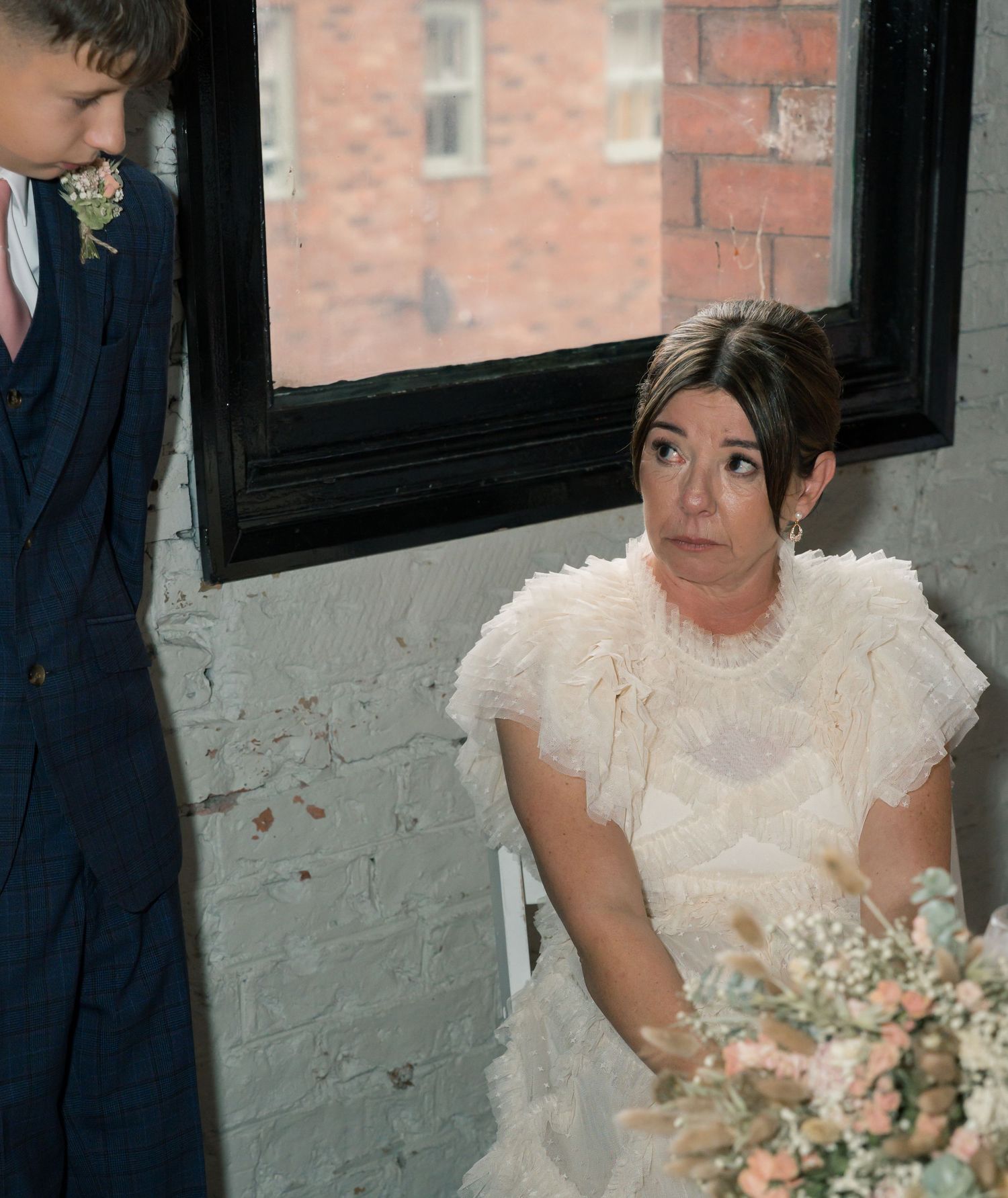 A bride in an elegant white ruffled dress poses with her bouquet against a brick wall backdrop.