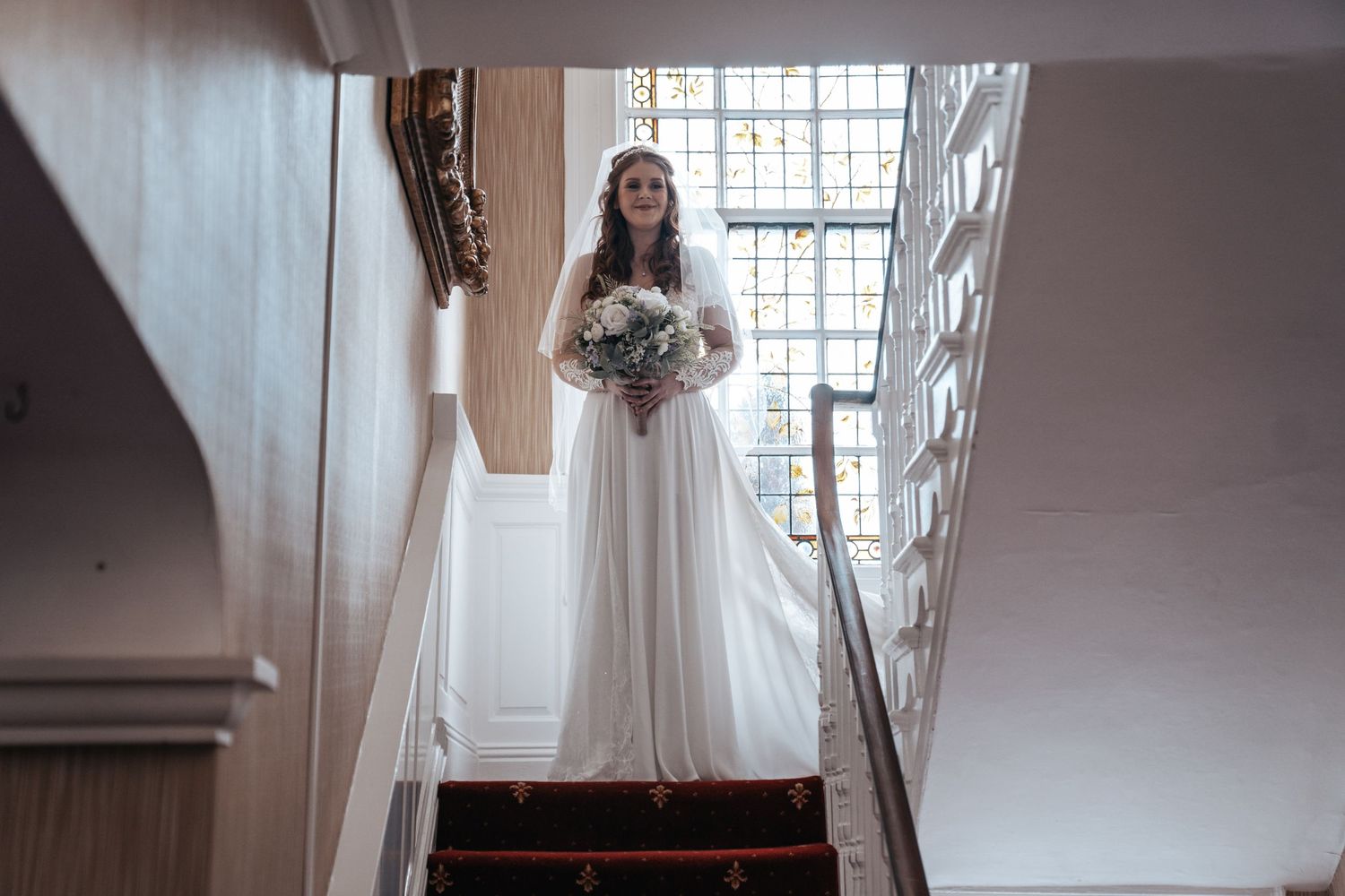 A white-gowned figure with bouquet stands gracefully at the top of a sunlit staircase.