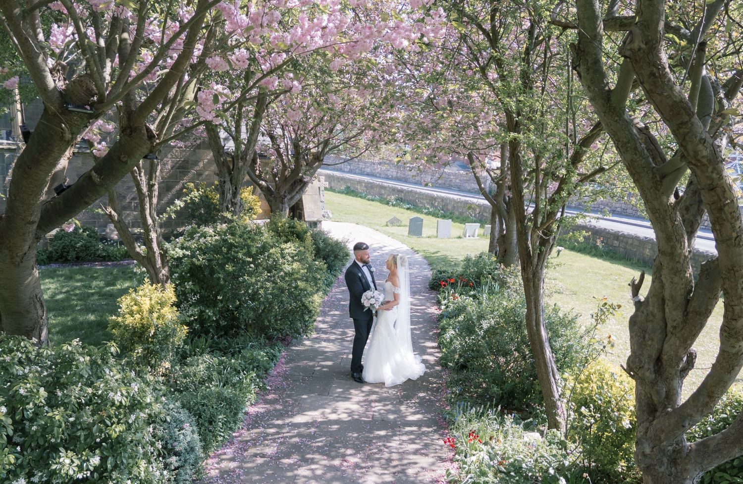 A bride and groom share a romantic moment on a garden path lined with blooming cherry blossom trees in spring, in Kippax Leeds.