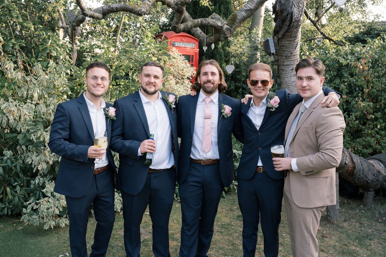 Wedding party members in dark suits share drinks and laughs outdoors.