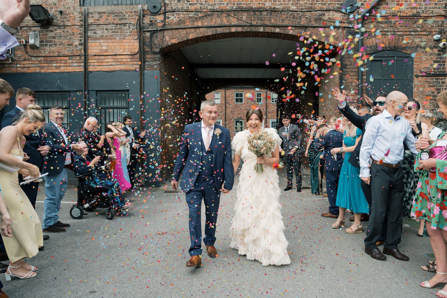 Wedding guests shower the newlyweds with colorful confetti outside a brick building during a joyful celebration.