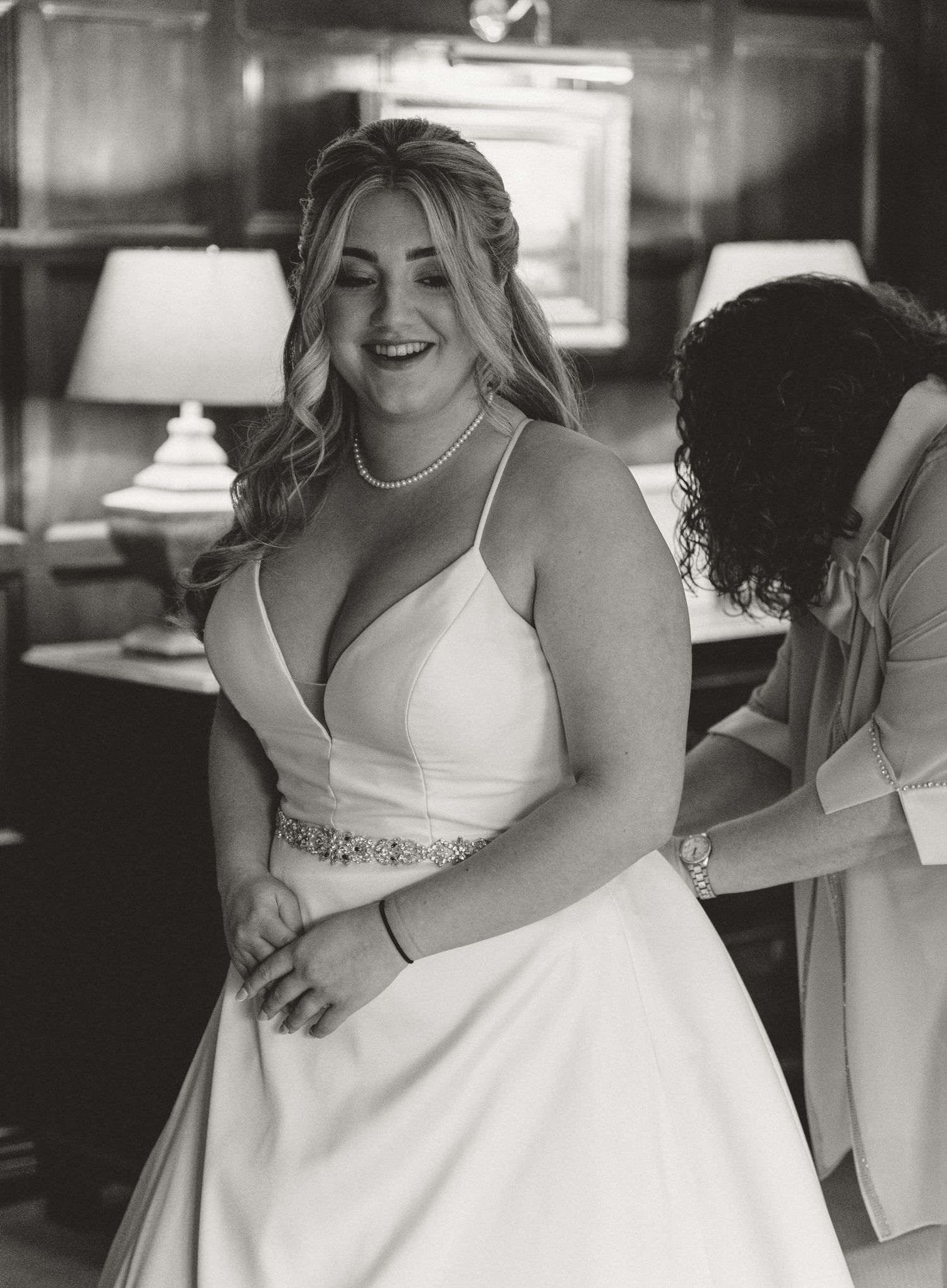 Black and white photo of a smiling bride in a white gown being helped into her dress by a bridesmaid at Whitley Hall.