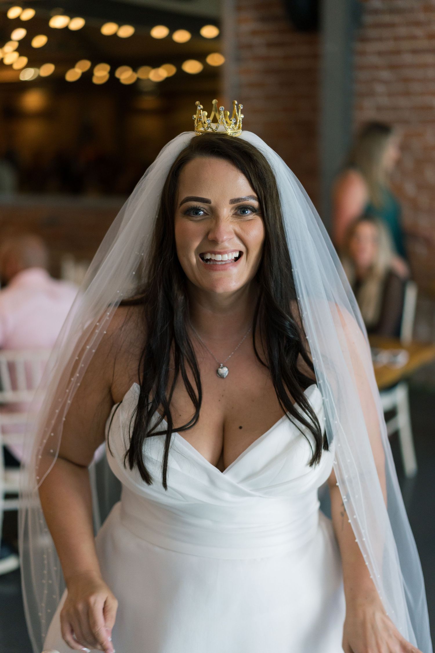 A bride in a white wedding dress and veil smiles during her wedding celebration with string lights in the background.
