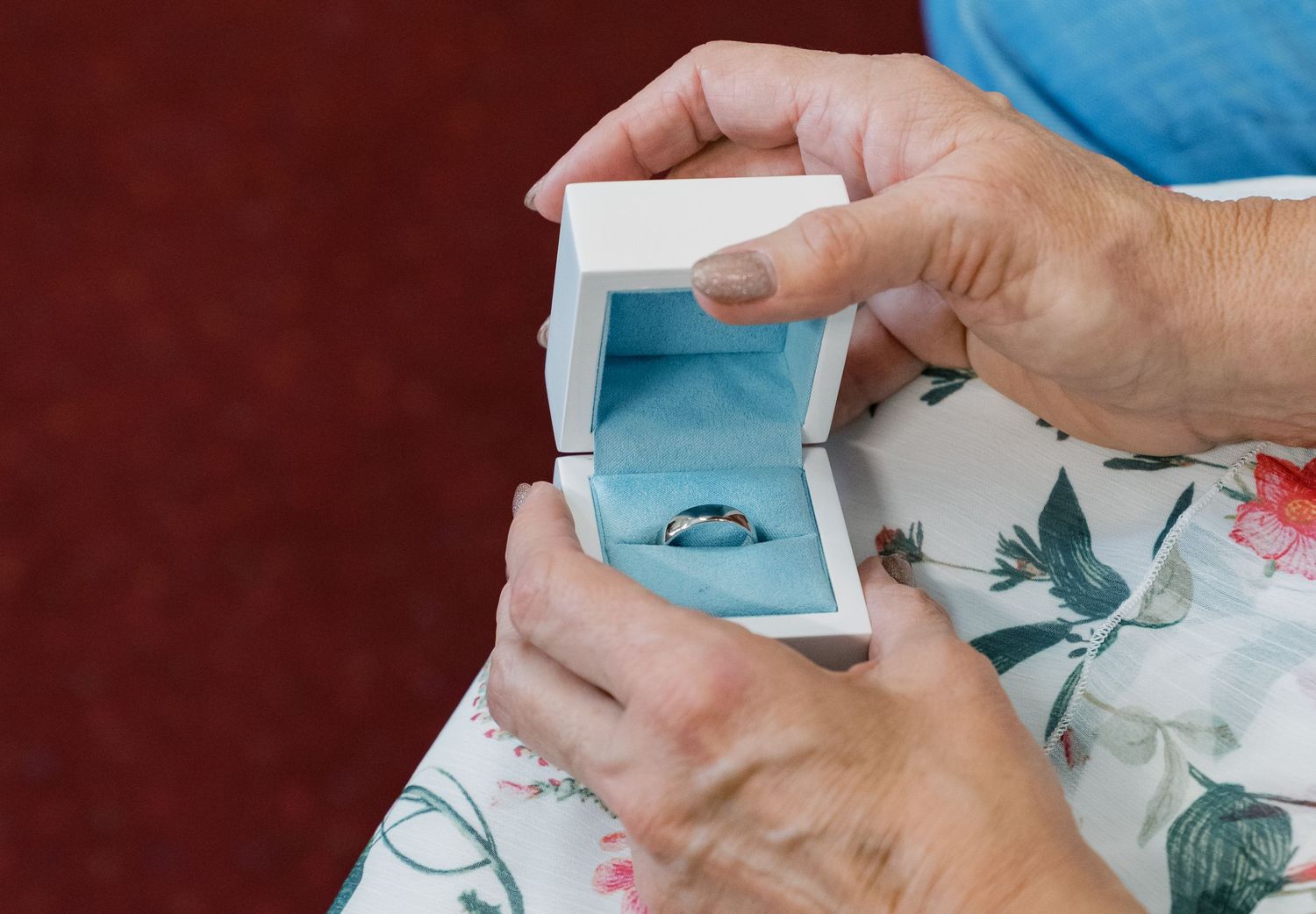 Hands opening light blue ring box containing wedding band against floral pattern.