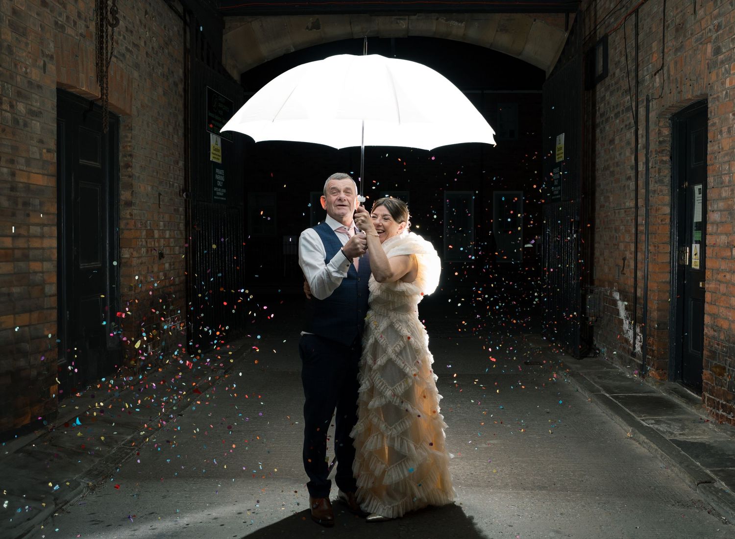 A wedding couple shares a romantic moment under an umbrella at night with confetti falling around them.