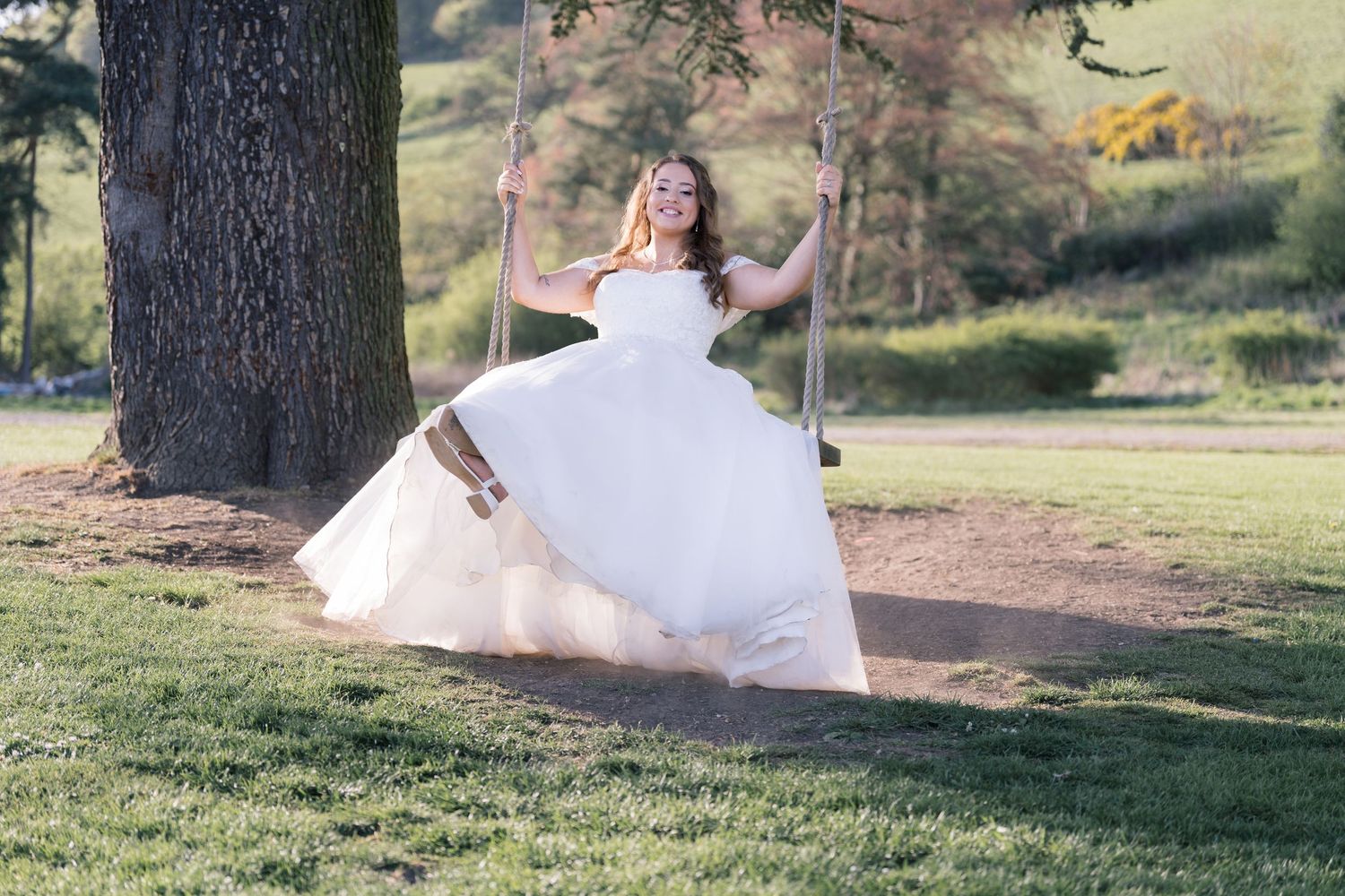 A person in a flowing white wedding dress swings joyfully on a tree swing in a scenic outdoor setting.