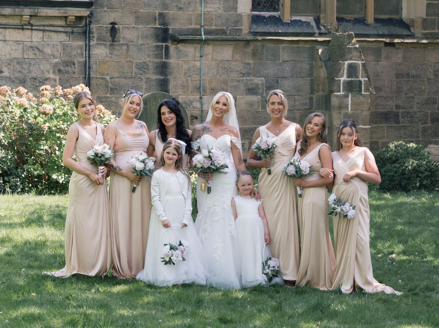 Bride in white gown poses with bridesmaids in blush pink dresses holding bouquets outside a stone church, in Leeds.