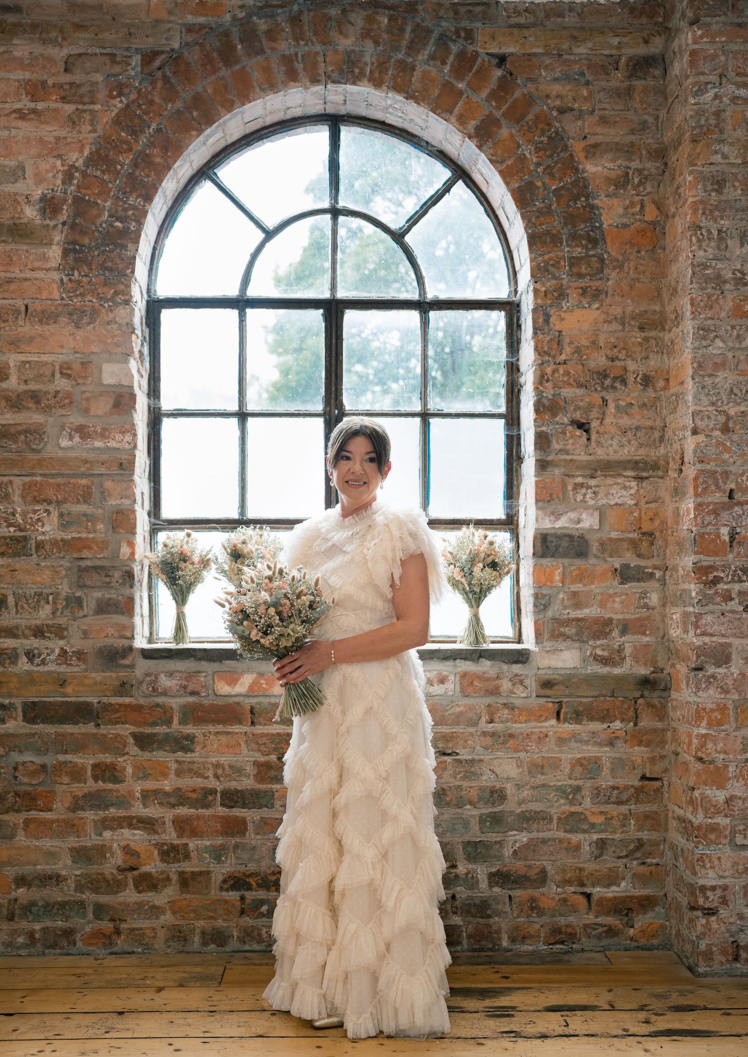 A bride in a ruffled white wedding gown poses in a rustic brick room with arched windows in a series of photos.