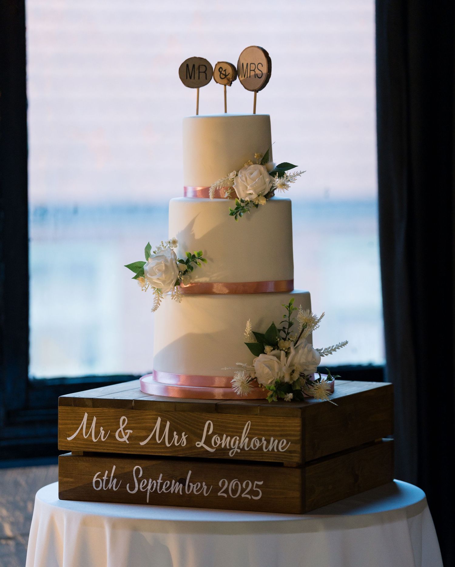 An elegant white wedding cake with floral decorations sits on a wooden display stand with custom text.