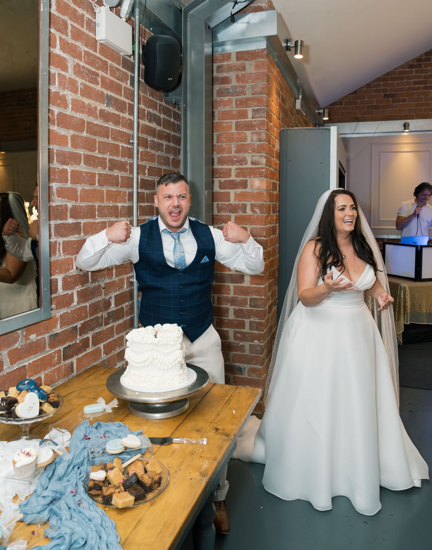 Wedding couple stands by their white tiered cake against an exposed brick wall at their reception.