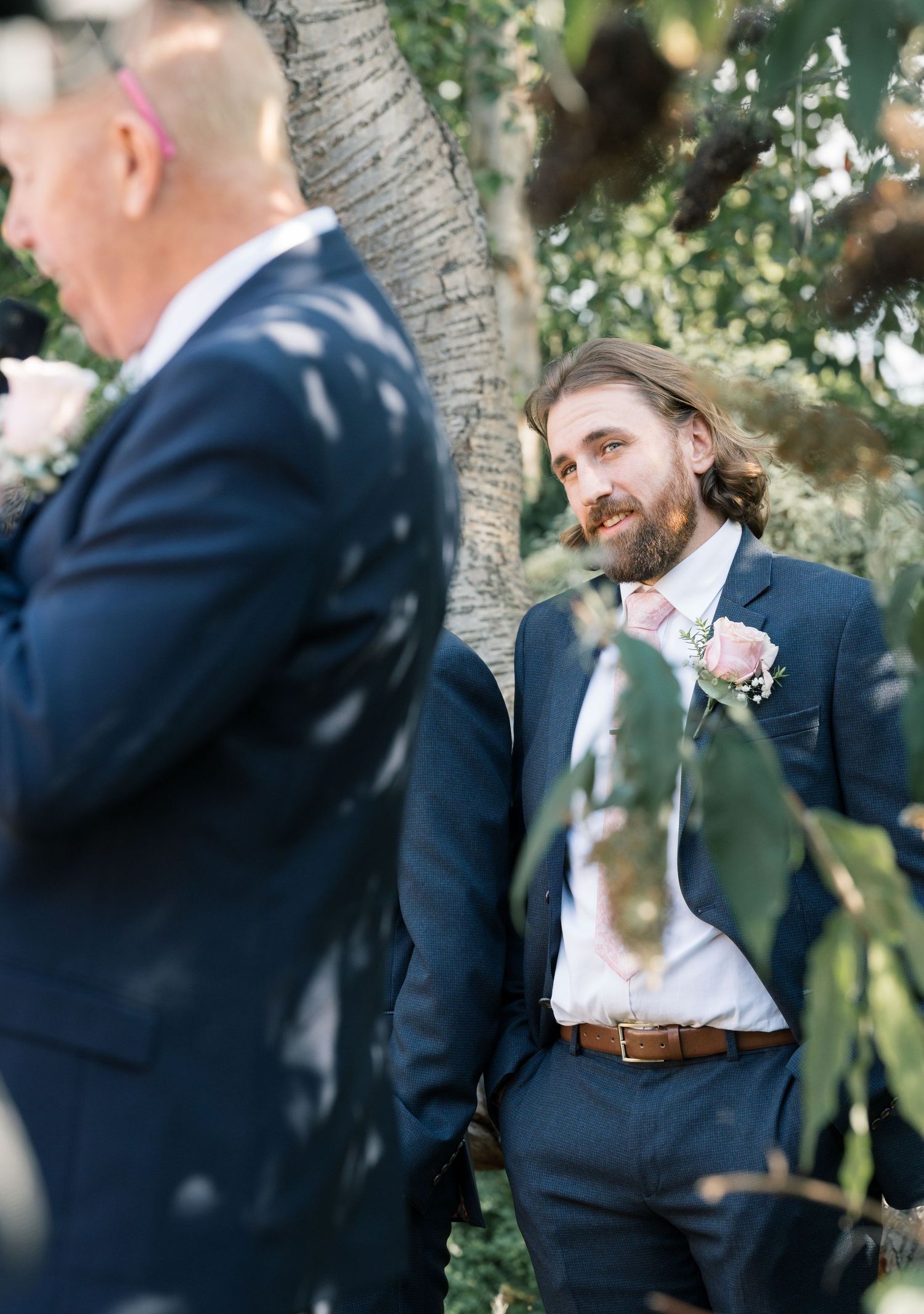 Wedding party members in navy suits gather under trees for outdoor photos.
