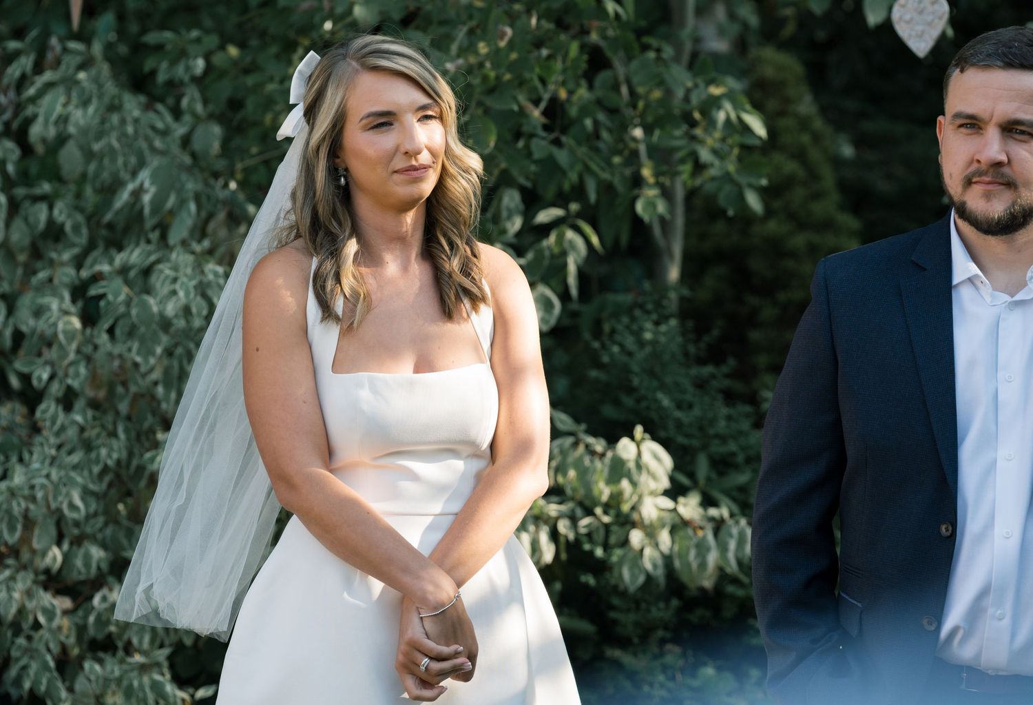 A bride in a strapless white wedding dress and veil stands next to a groom in a navy suit during an outdoor ceremony.