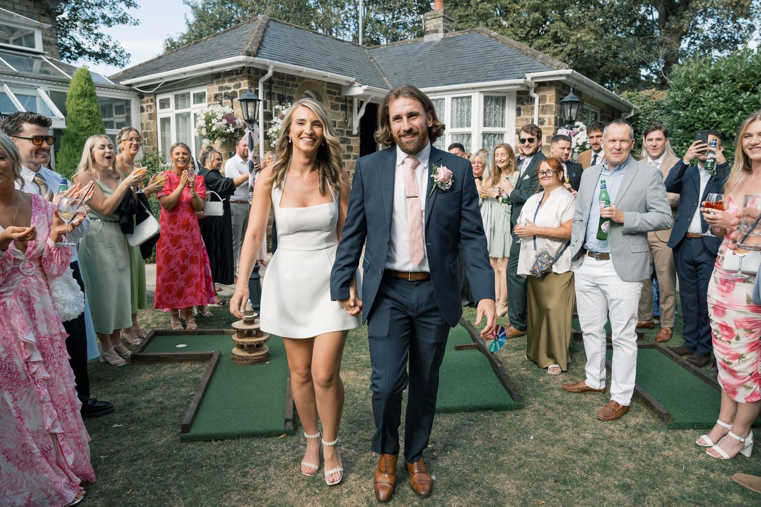 Wedding guests form a celebratory receiving line outdoors on a sunny day to welcome the newlywed couple.